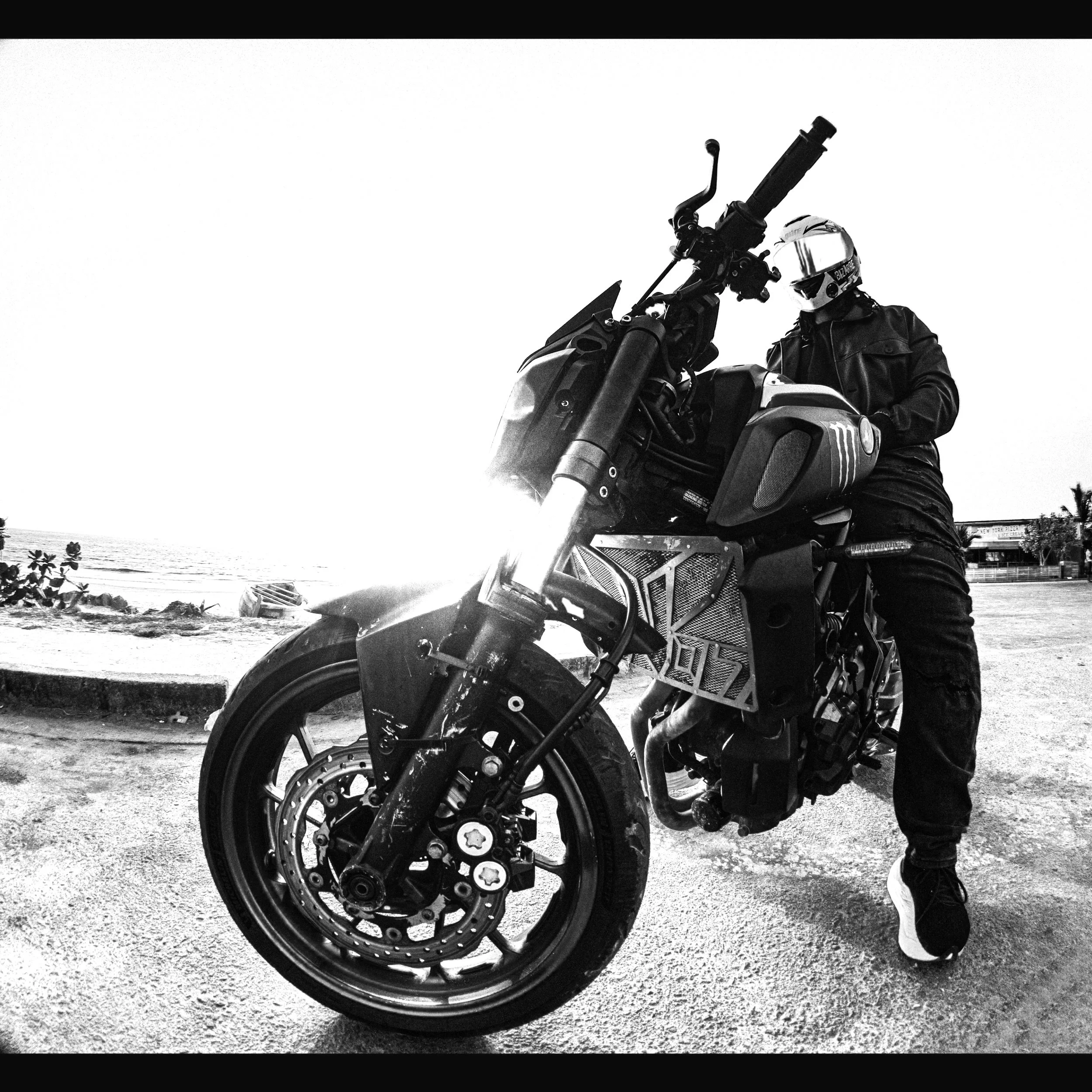 A person in black riding gear and a colorful helmet beside a black motorcycle on a beach at sunset.