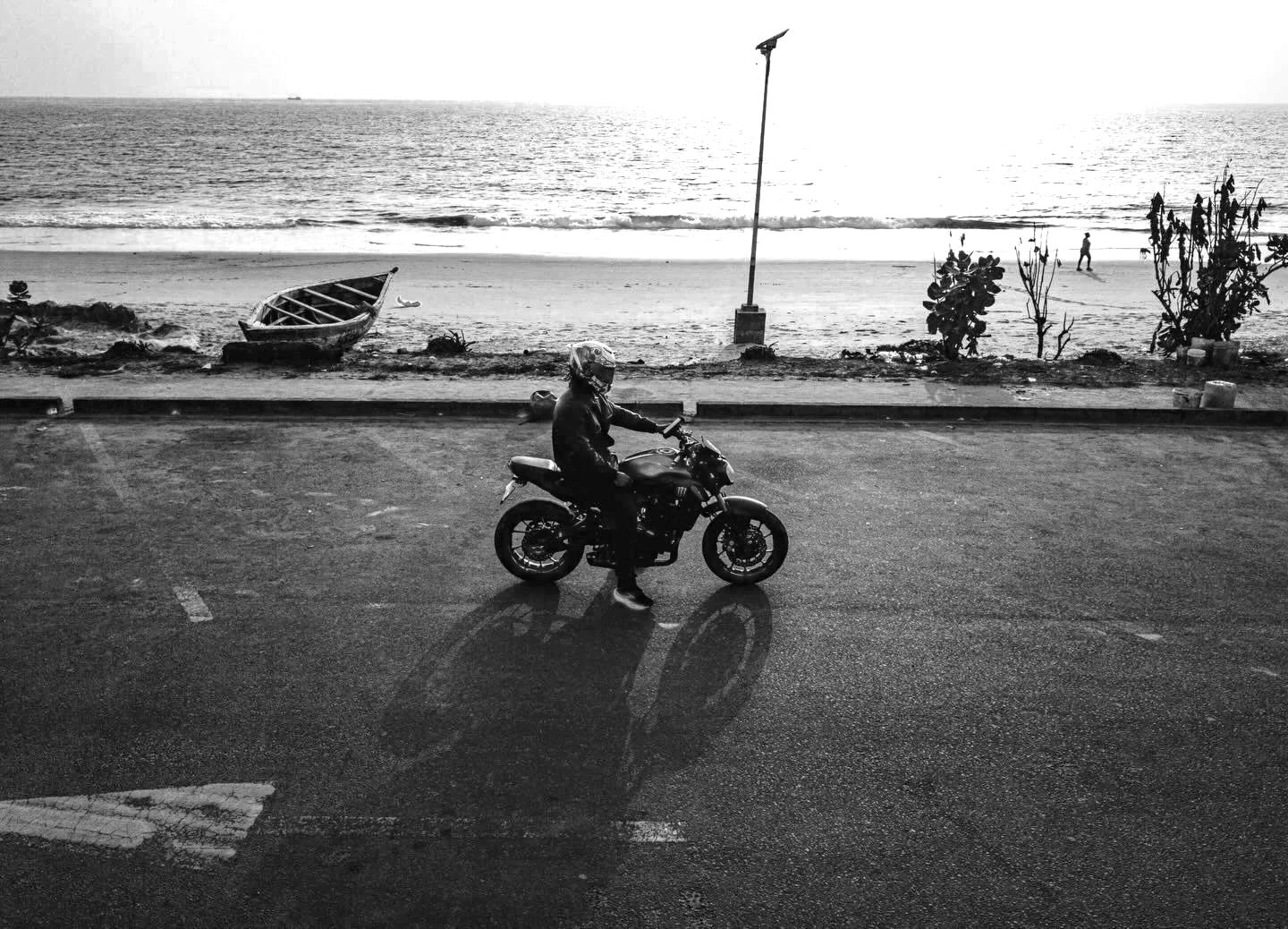 A person riding a motorcycle on a paved road near the beach, with an overturned boat, bushes, and the ocean in the background, during sunset or sunrise.