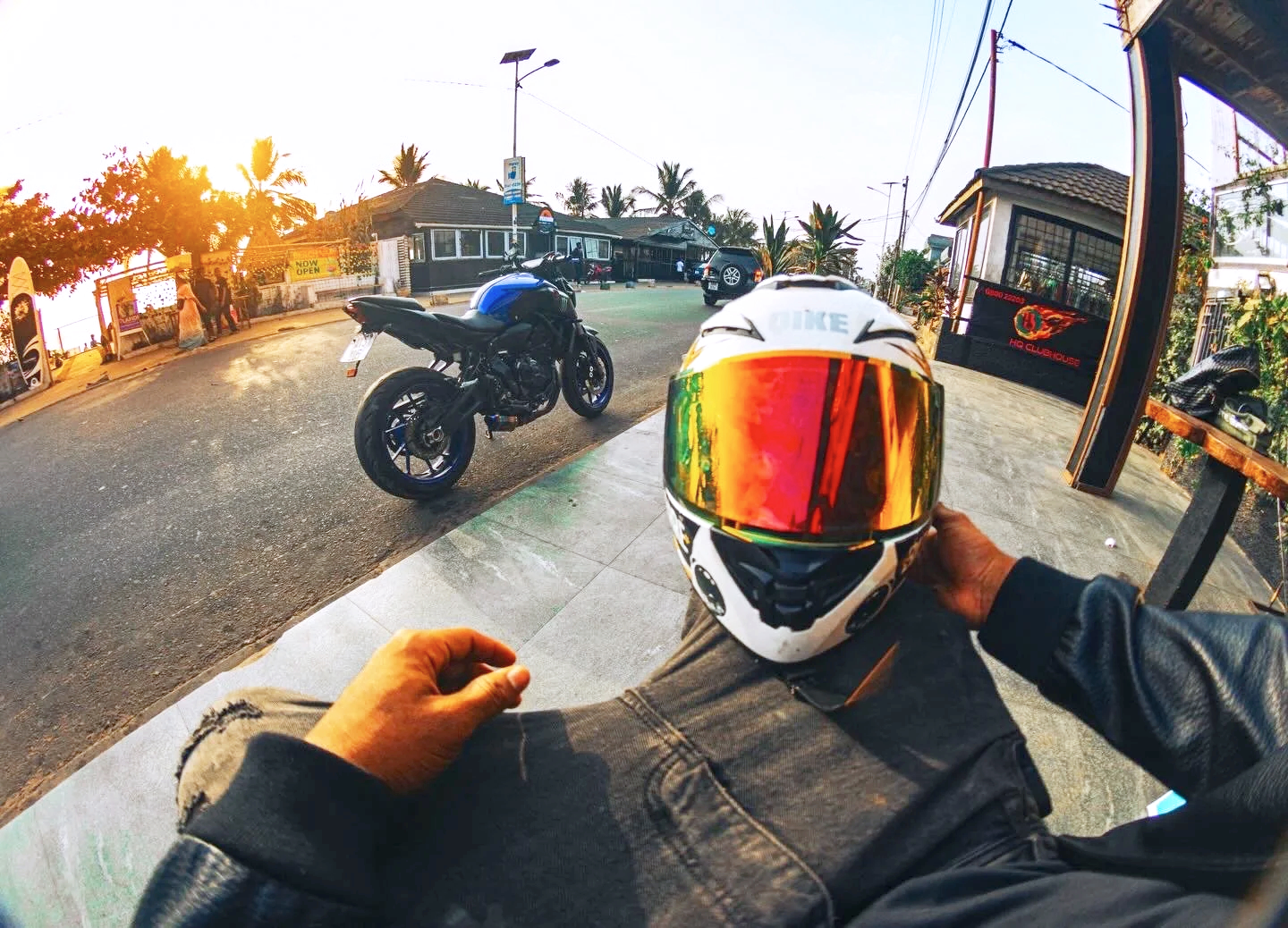 Person sitting on a motorcycle holding a white helmet with a reflective red visor, another motorcycle parked on the street, tropical trees and buildings in the background at sunset.