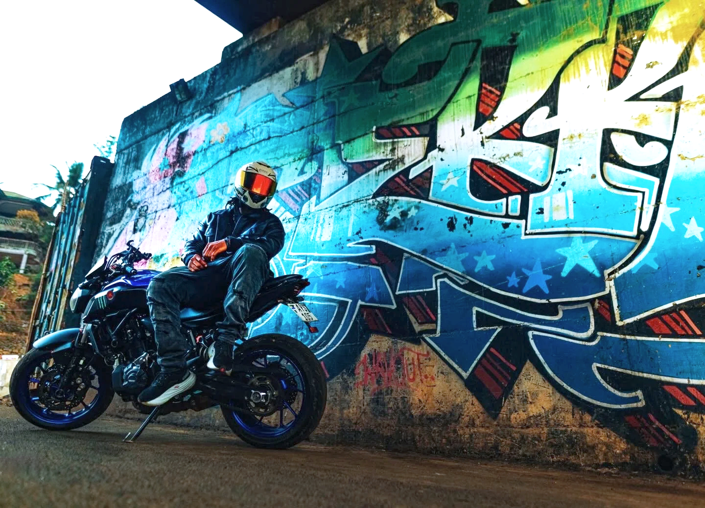 Person wearing a helmet and leather jacket sitting on a motorcycle in front of a vibrant graffiti wall.