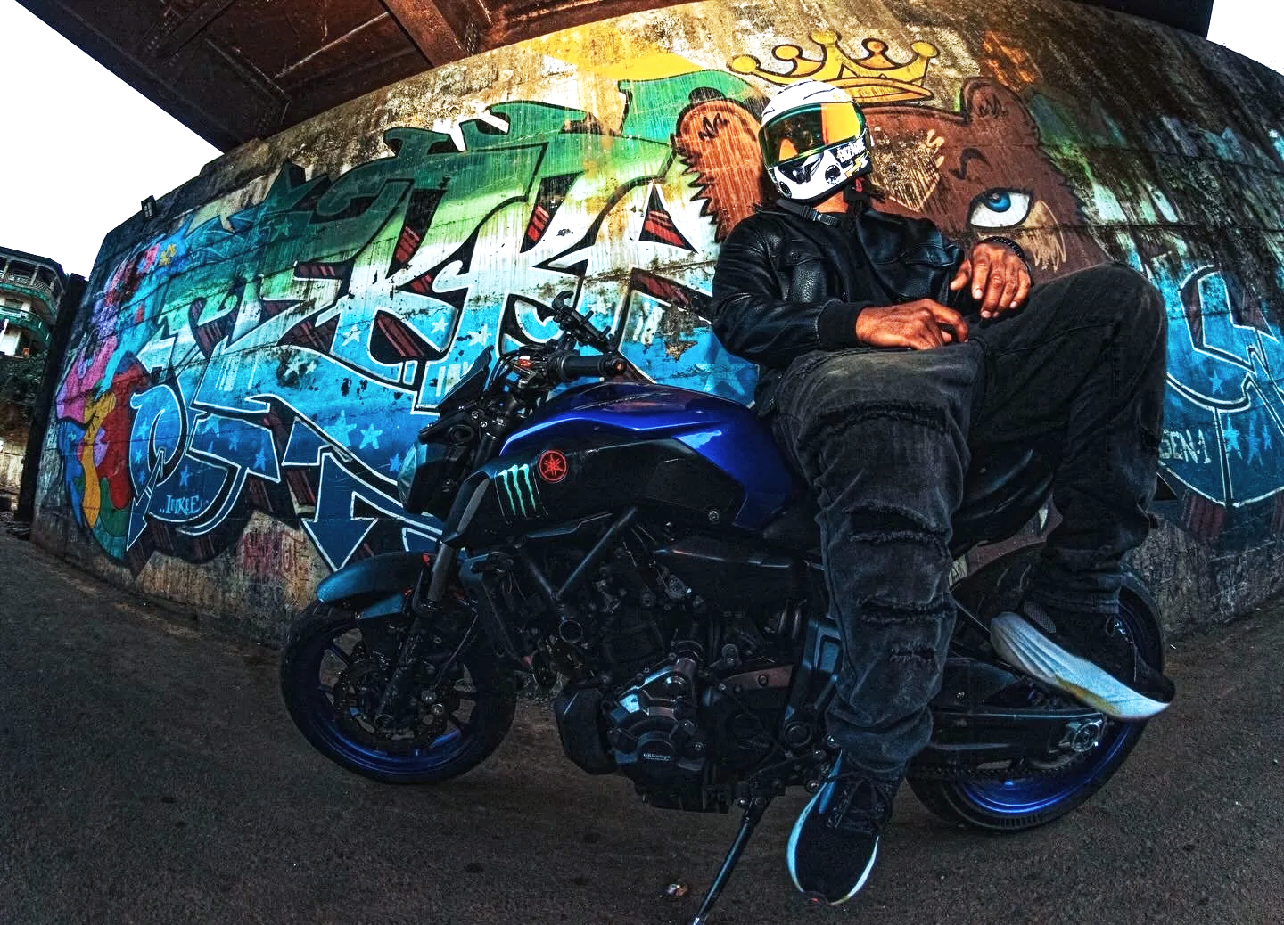 A man in black motorcycle gear and a helmet sitting on a blue motorcycle, with a colorful graffiti mural on the wall behind him.