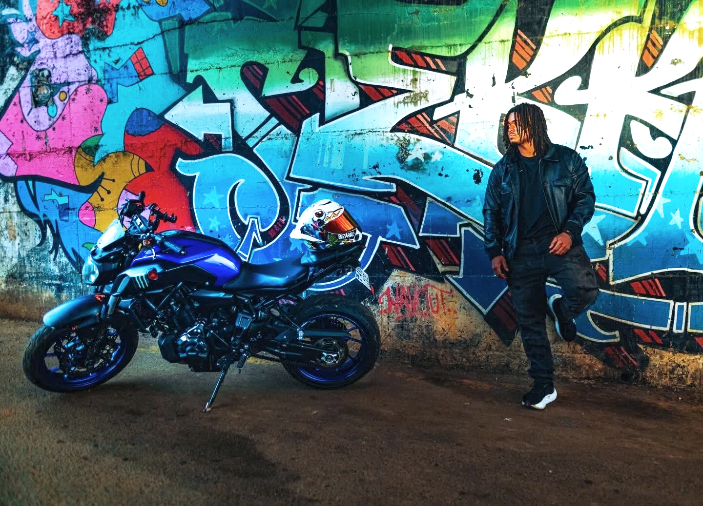 A person with dreadlocks wearing a black leather jacket and black pants leaning against a wall with colorful graffiti art. A blue motorcycle is parked beside them with a white helmet on the seat.