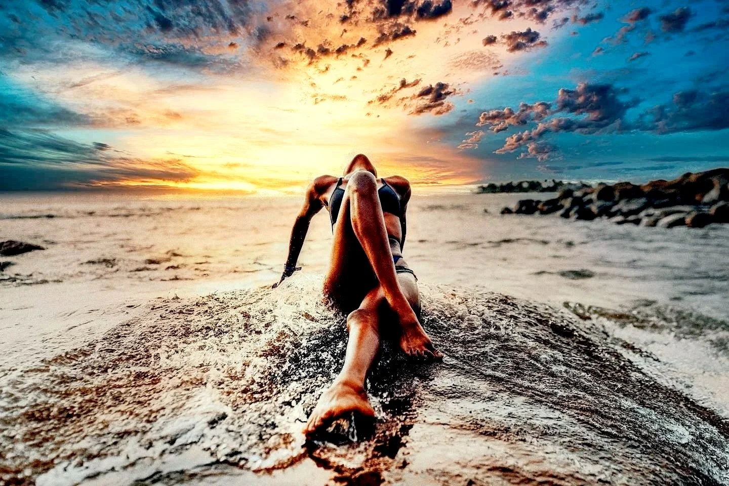A woman in a bikini reclining on a surfboard on the beach during a sunset, with a colorful sky and clouds in the background.