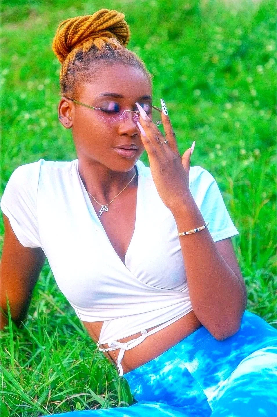 A woman with blonde braids styled in a bun listens through glasses with decorative elements, sitting on grass in a park, wearing a white wrap-top and blue tie-dye pants, accessorized with jewelry.