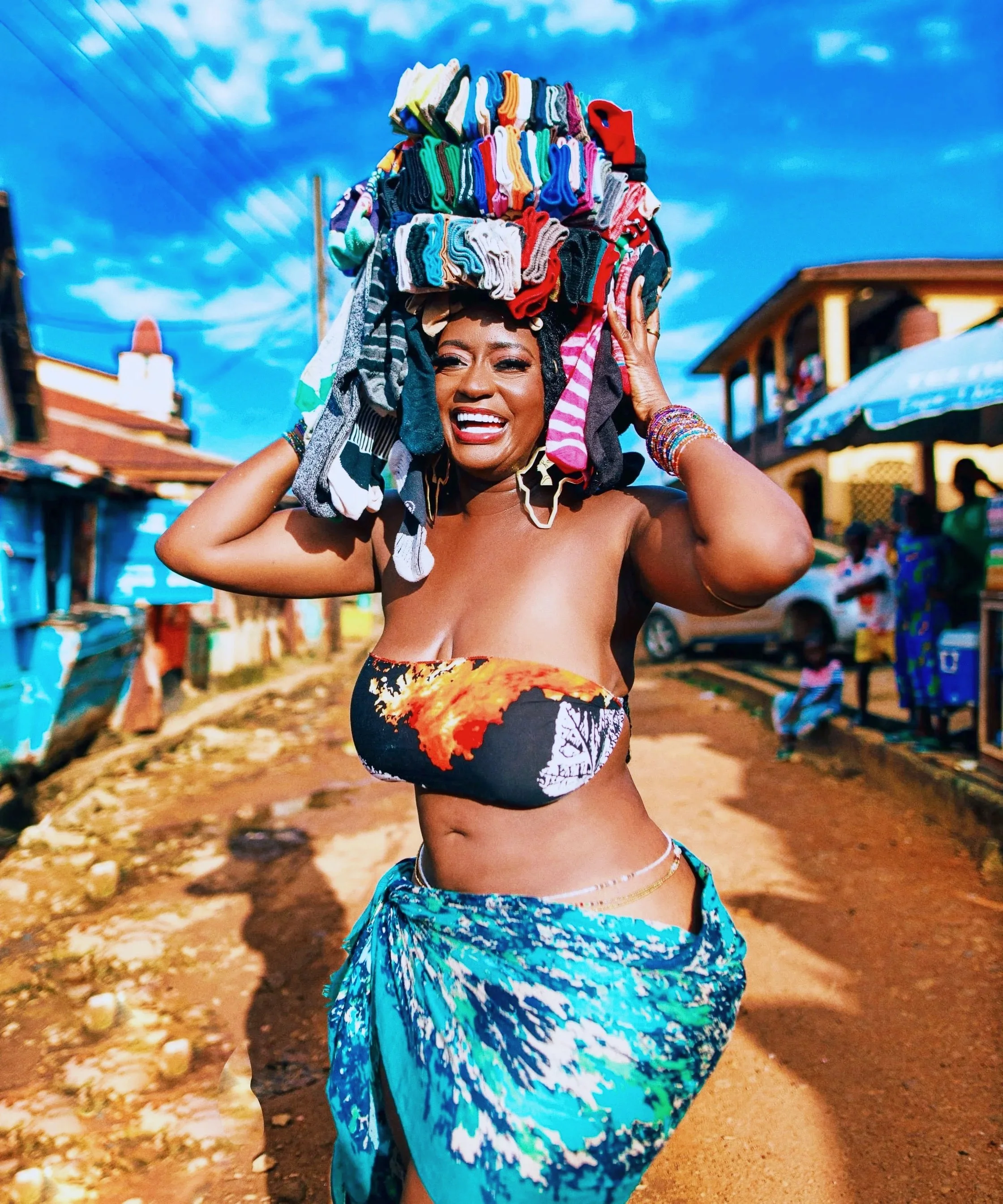 A woman smiling with a large basket of colorful socks balanced on her head, standing outdoors on a dirt road with buildings and people in the background on a sunny day.