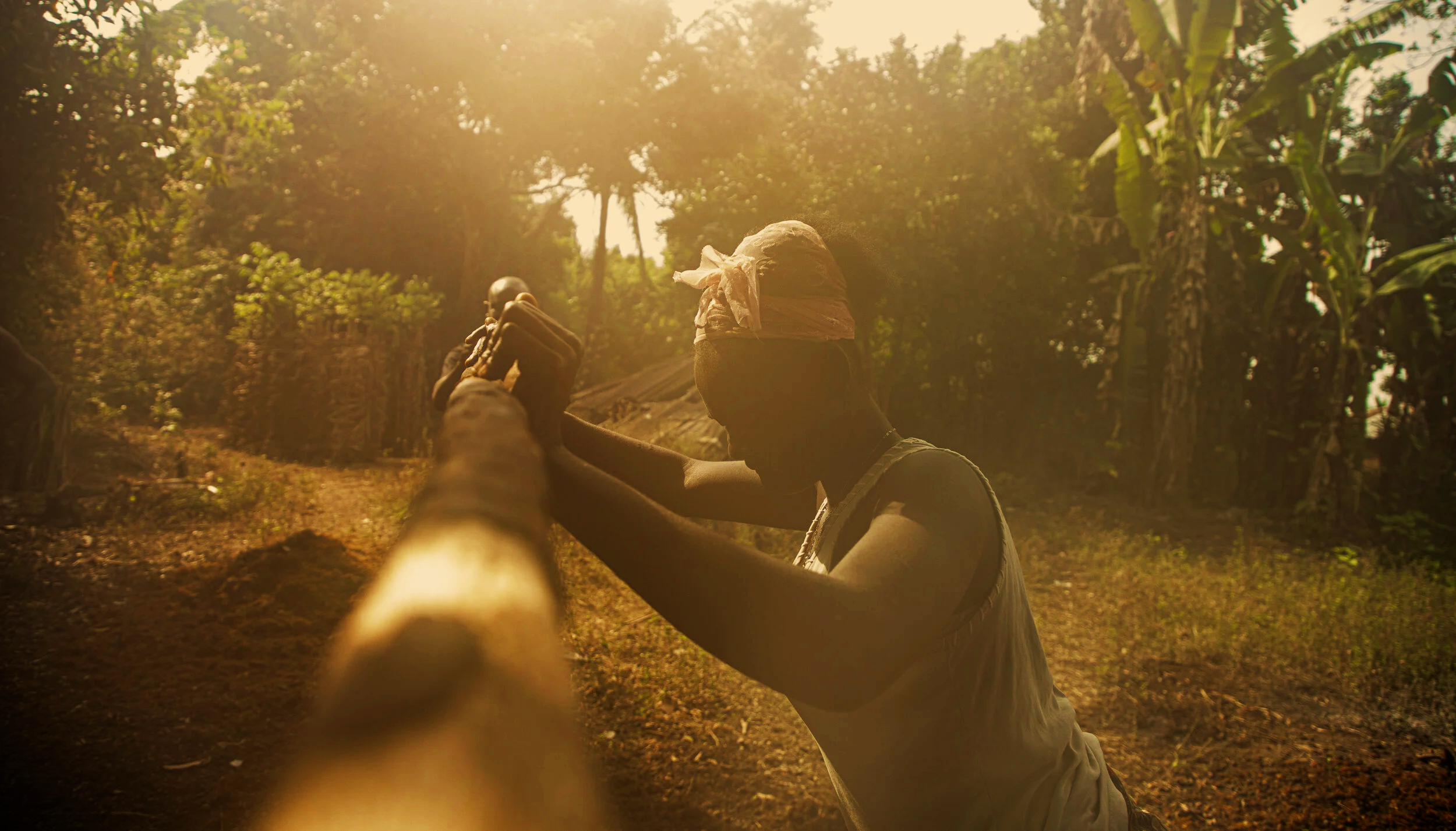 A young person with a headband and sleeveless shirt pulling a rope with a small wooden hut and lush green trees in the background, sunlight shining through the trees.