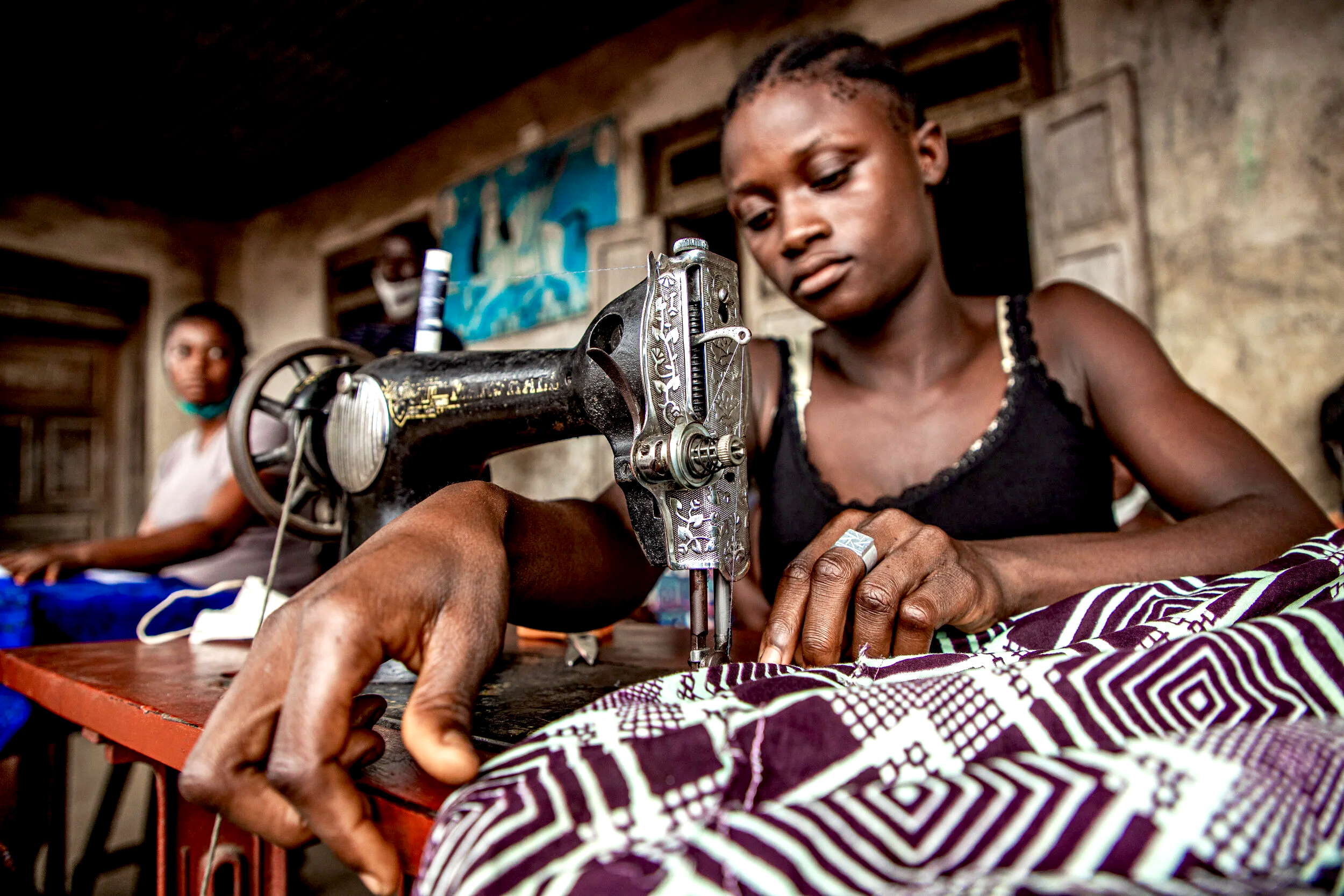 A young woman sewing fabric on an old-fashioned sewing machine inside a rustic room with two other women in the background.