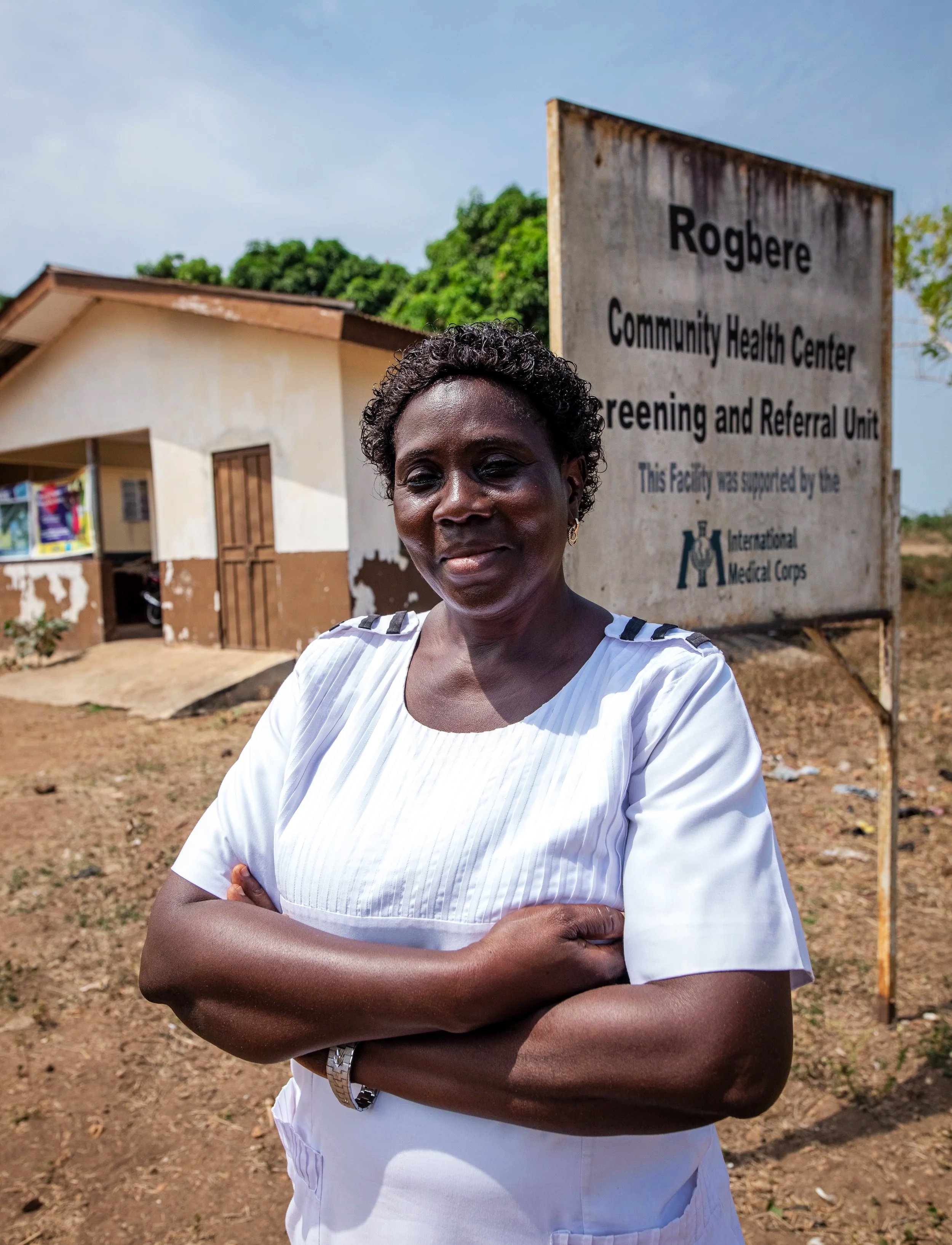A woman in white medical scrubs standing outdoors with arms crossed, in front of a sign for Rogbere Community Health Center in a rural setting.