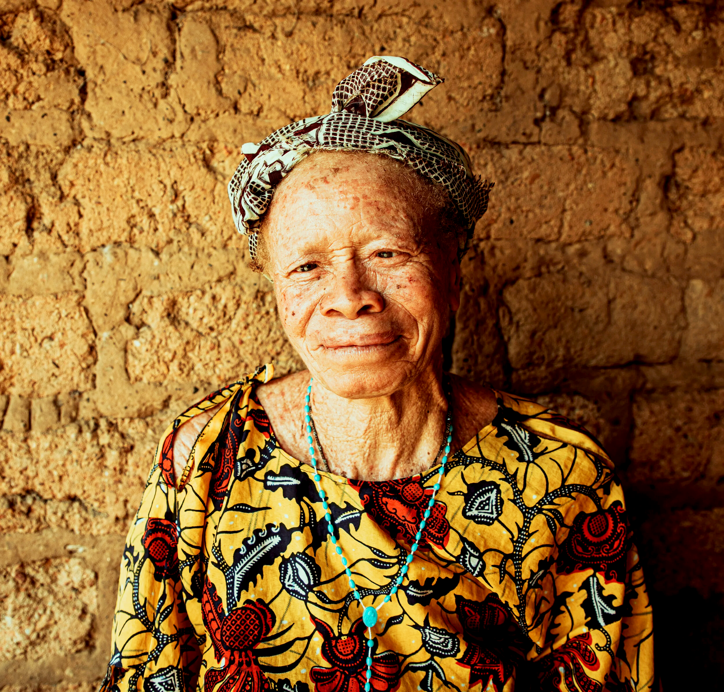 An elderly woman with a warm smile, wearing a patterned headscarf, a colorful printed dress with red and black designs, and a turquoise beaded necklace, standing in front of a textured mud wall.