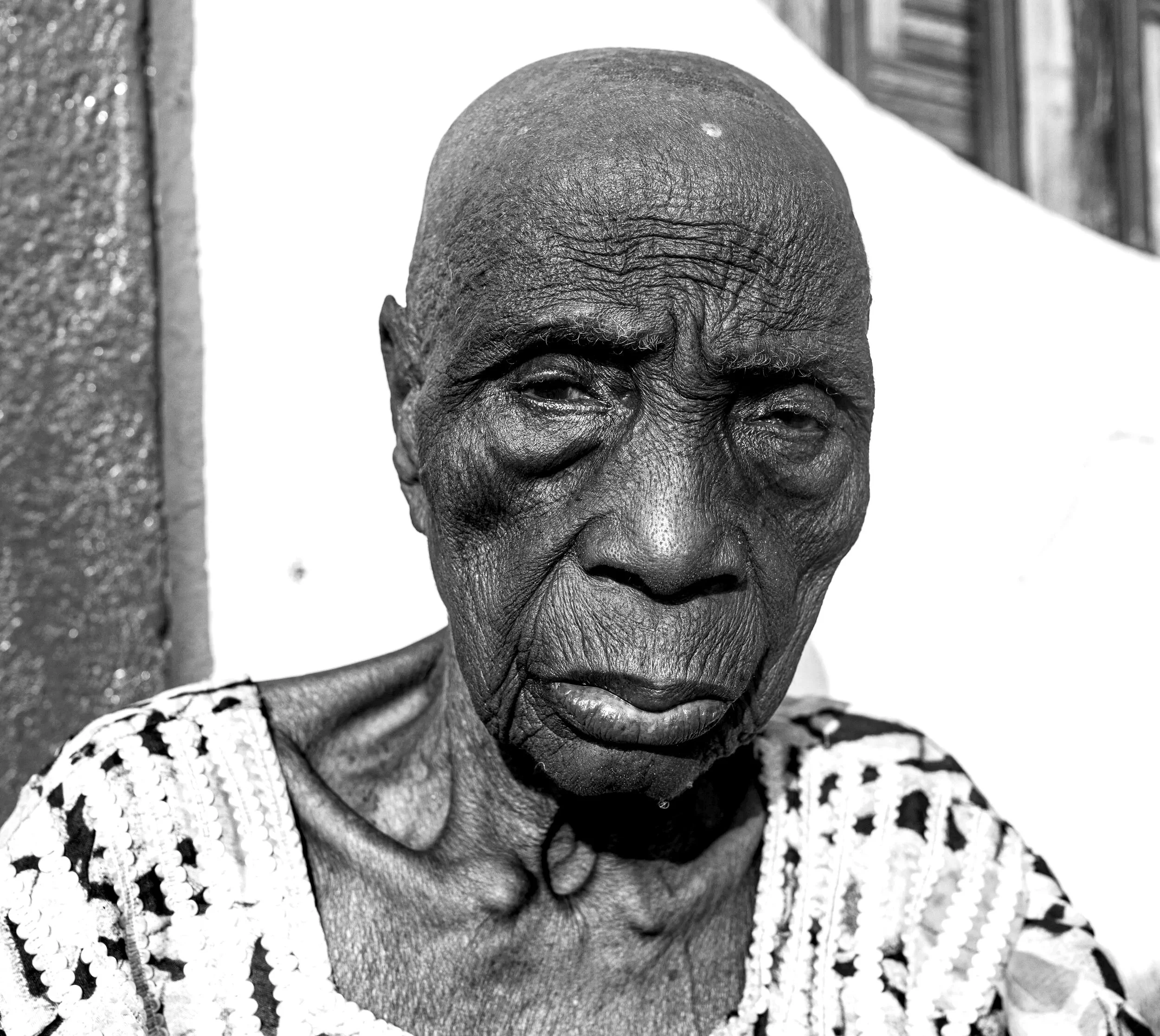 Close-up black and white portrait of an elderly woman with deep wrinkles, wearing a patterned top, standing outdoors near a wall.