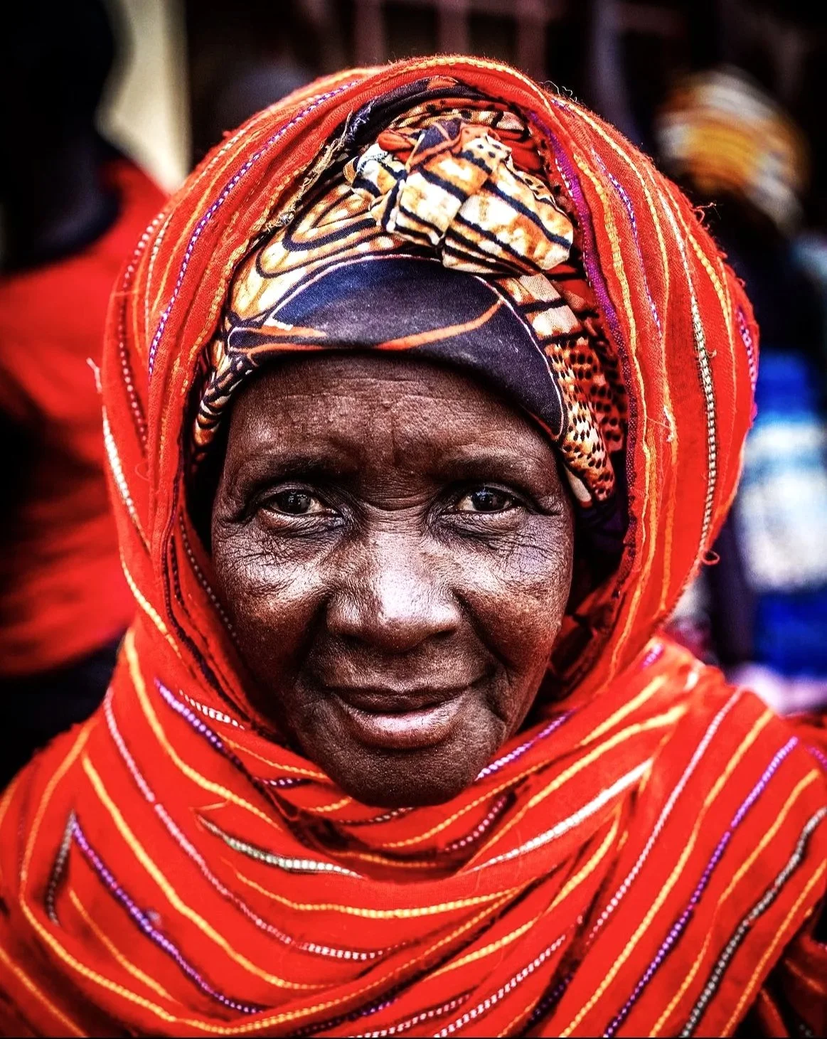 Close-up of an elderly woman with dark skin, wearing a vibrant red patterned head wrap and shawl, smiling softly. Background shows blurred people and warm lighting.