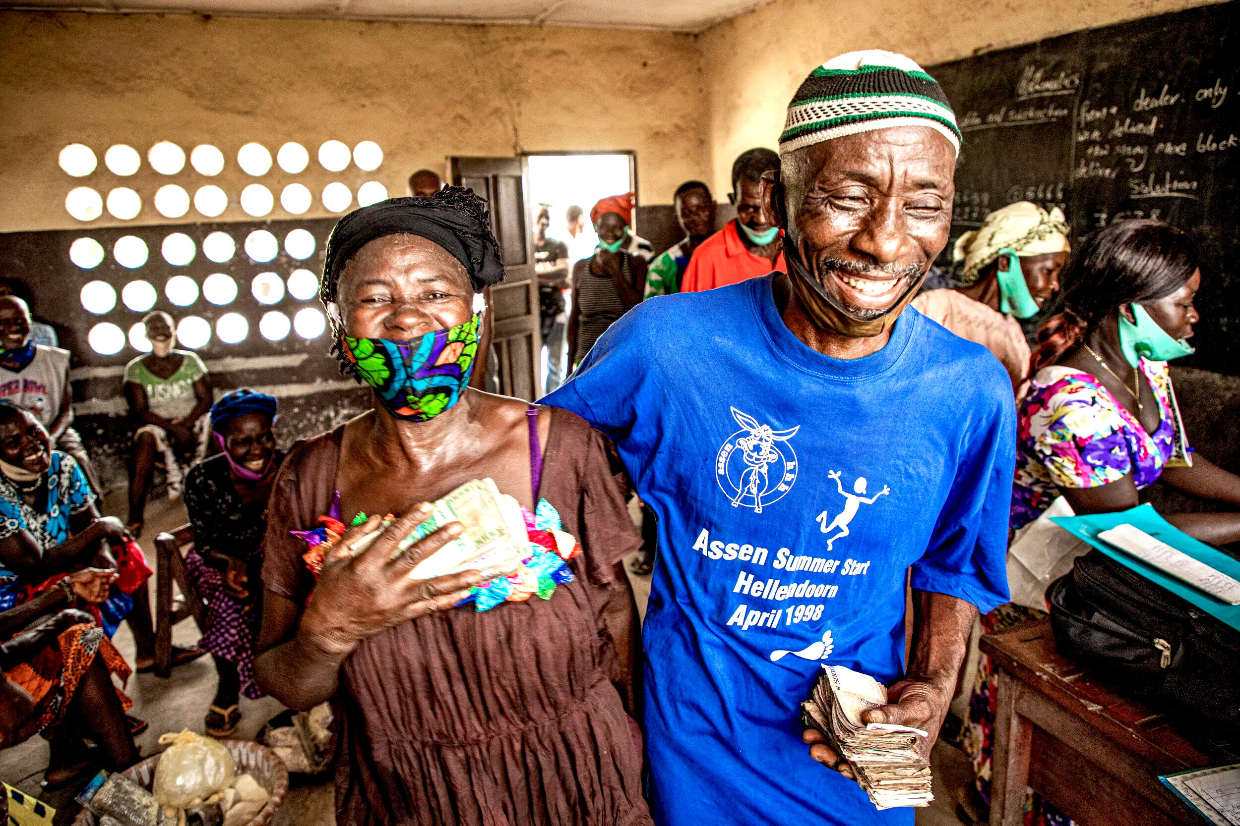 A smiling elderly man holding a stack of money, standing beside an elderly woman who is also smiling and holding some money close to her chest. They are in a room filled with other smiling people, some wearing face masks, with a blackboard and a wind