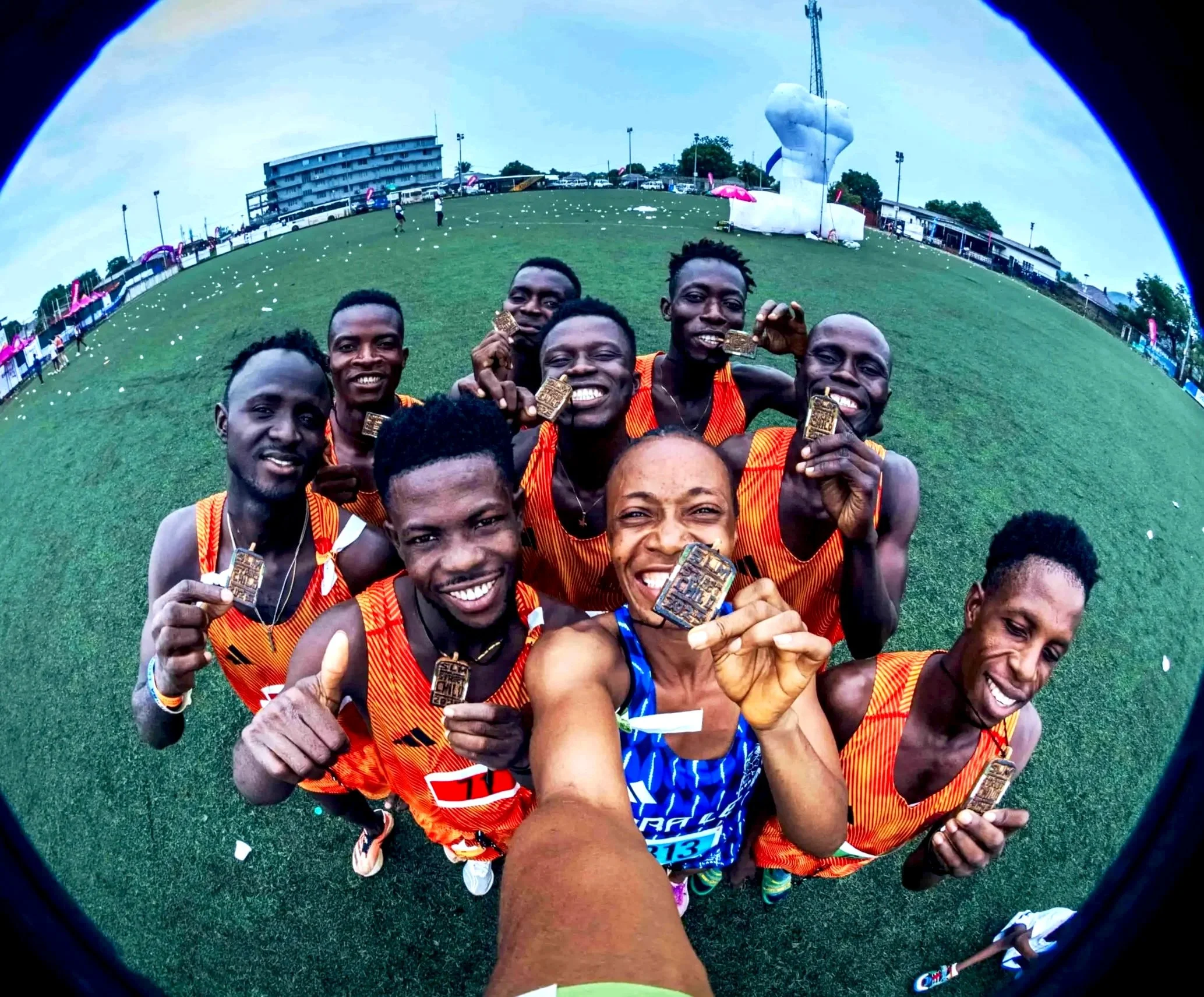 Group of smiling runners holding medals in a grassy field after a race, with a large white sculpture and event tents in the background.