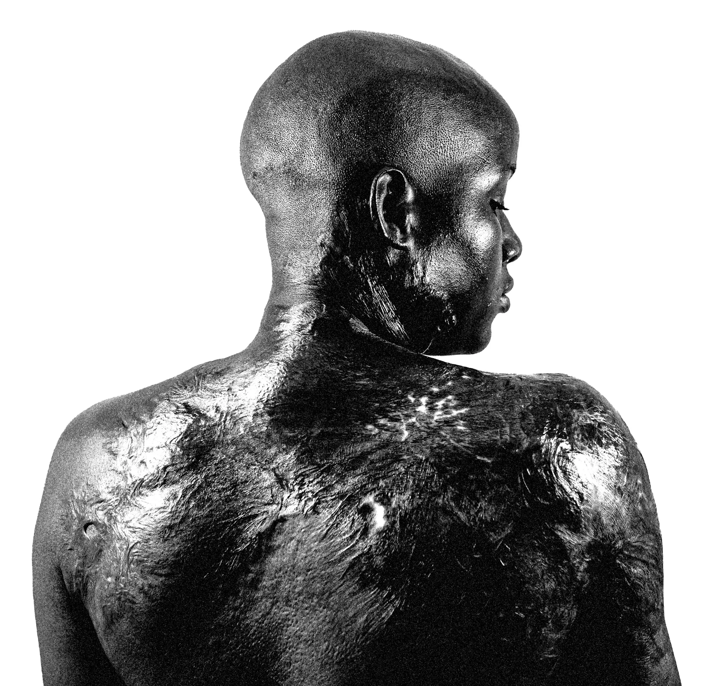 Black woman with short hair, turned to the side, showing her shoulder and back covered in scrasl, against a plain white background.