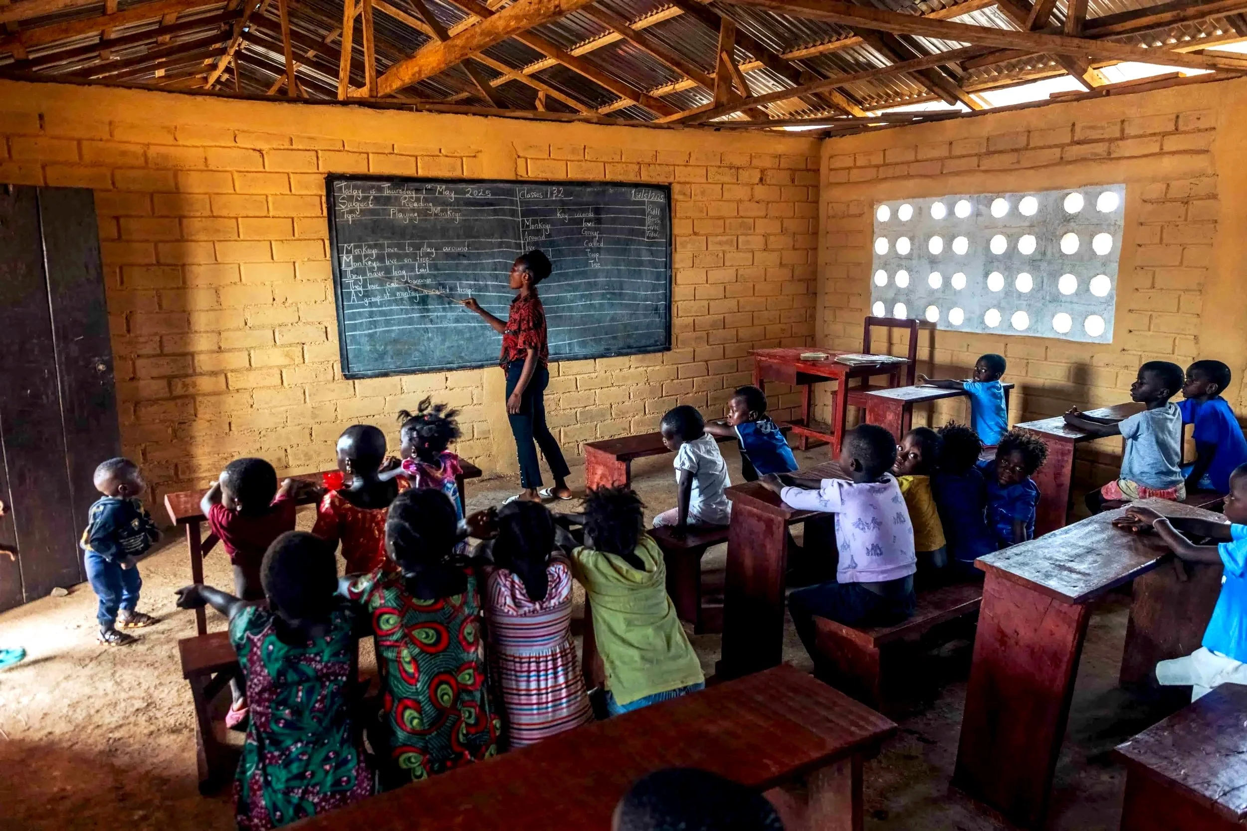 Classroom with children sitting at desks and a teacher or student standing at blackboard with writing. The classroom has brick walls and a partially open window with circular holes in the concrete.