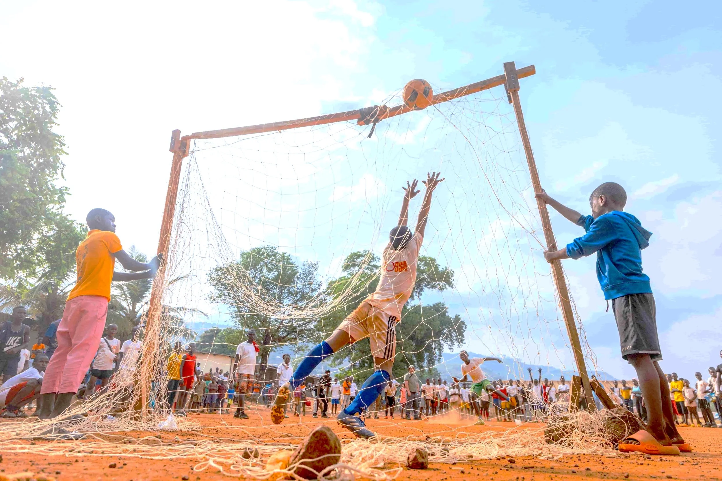 Children playing soccer in a dirt field with a crowd watching, the picture taken from the ground level showing a soccer goal and players jumping to reach the ball.