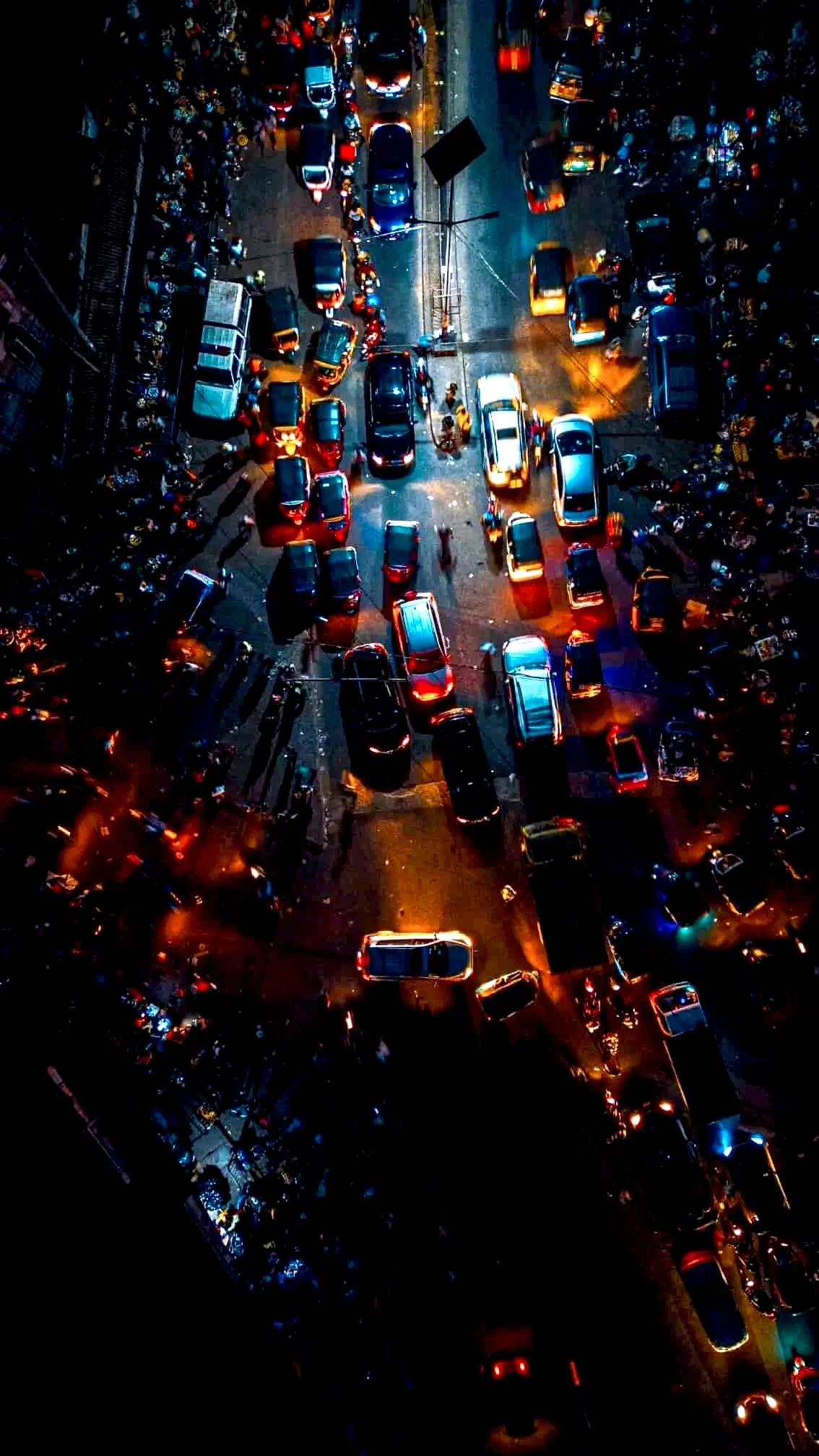 An aerial nighttime view of a busy city street filled with cars, motorcycles, and people, illuminated by streetlights and vehicle headlights.