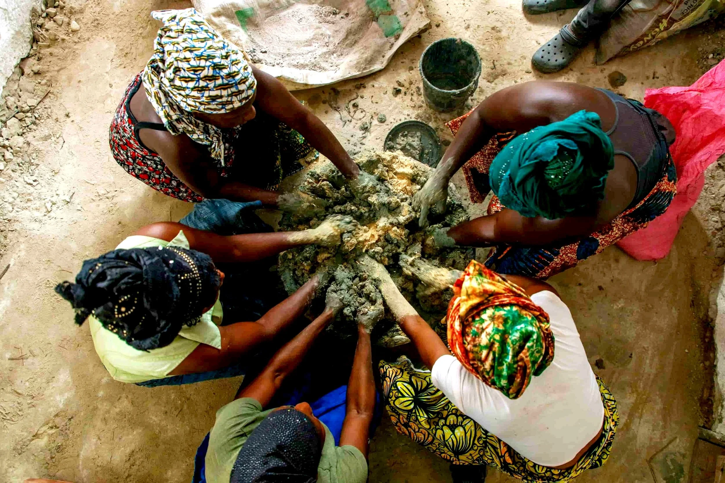 Several women are gathered around a pile of mud or clay, working together in a communal activity. They are outdoors on a dirt ground, wearing colorful clothing and headscarves, and appear to be engaged in a traditional craft or construction task.