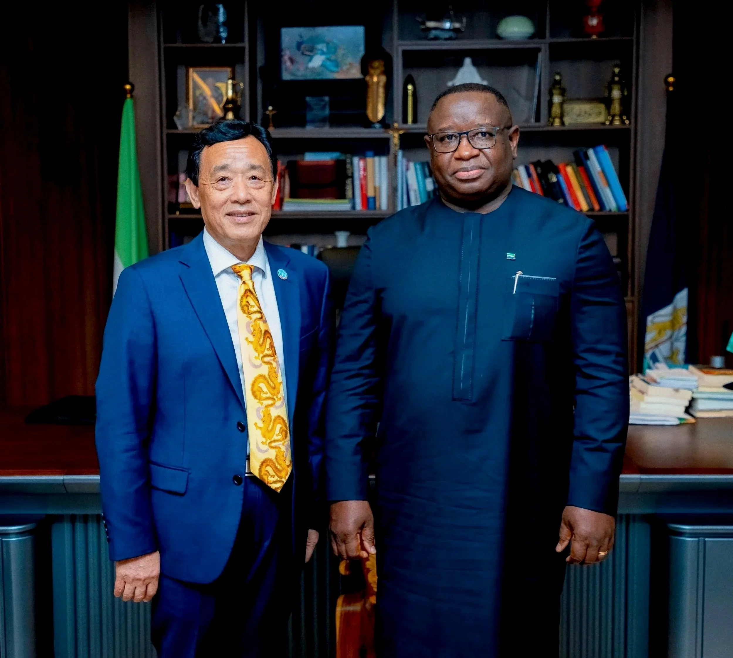 Two men standing in front of a bookshelf with various decorative items and books, holding hands.