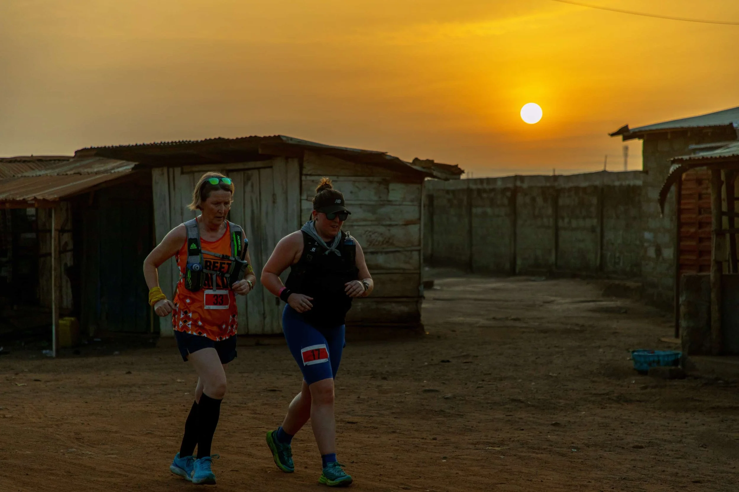 Two women jogging on a dirt road during sunset, with rustic wooden houses in the background.