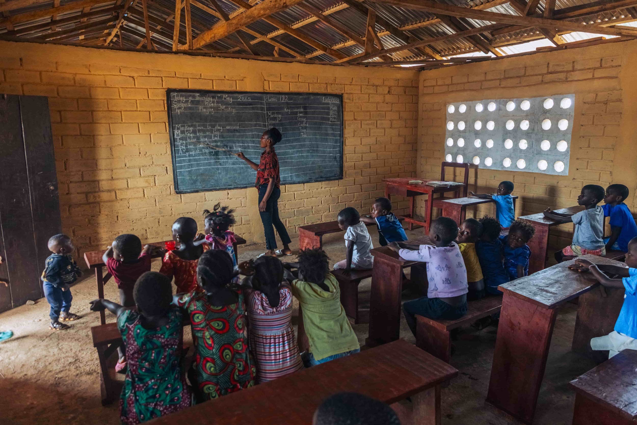 Classroom with children sitting at desks and a teacher or student standing at blackboard with writing. The classroom has brick walls and a partially open window with circular holes in the concrete.
