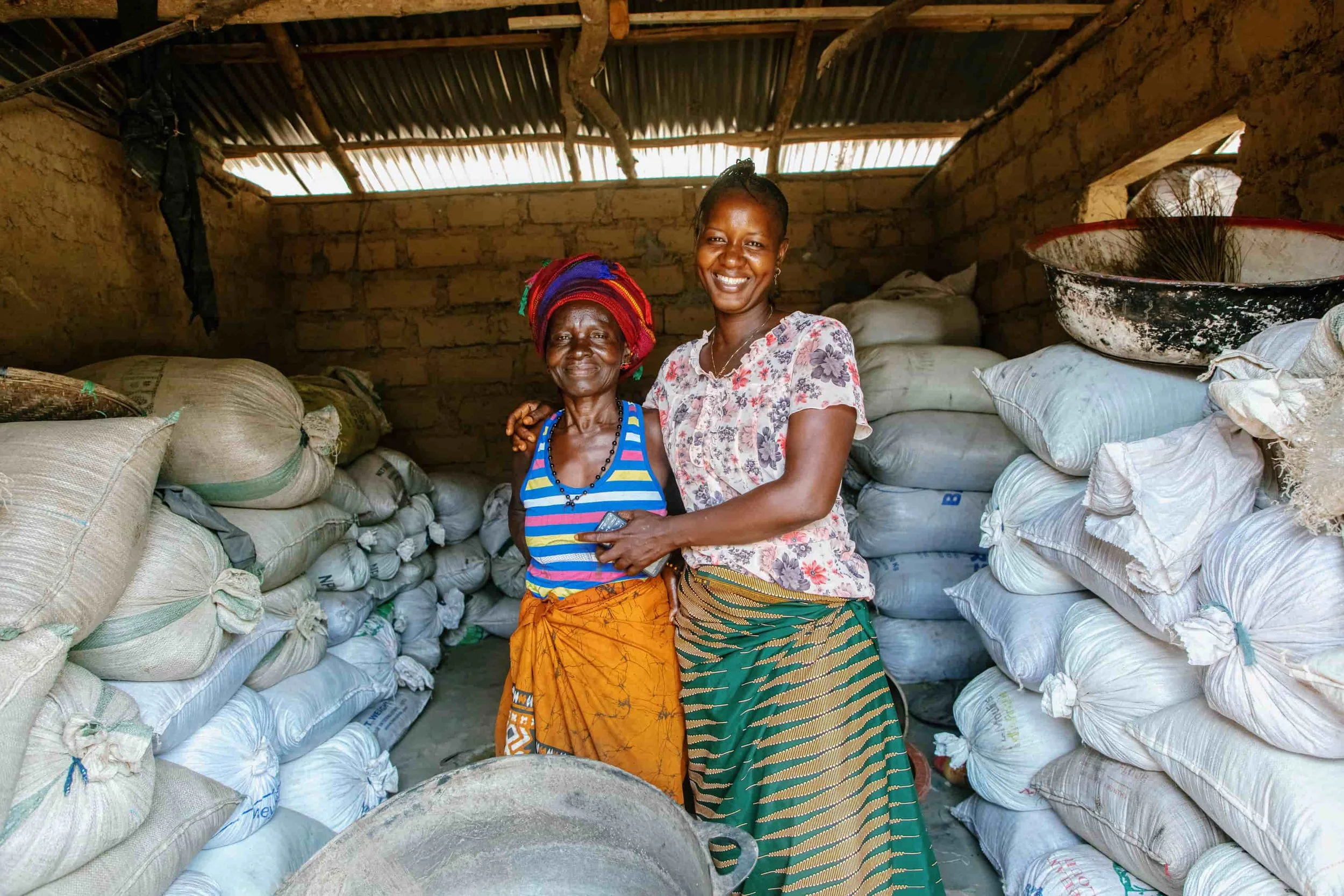 Two women standing inside a storage room filled with large bags of materials, smiling and posing for the camera.