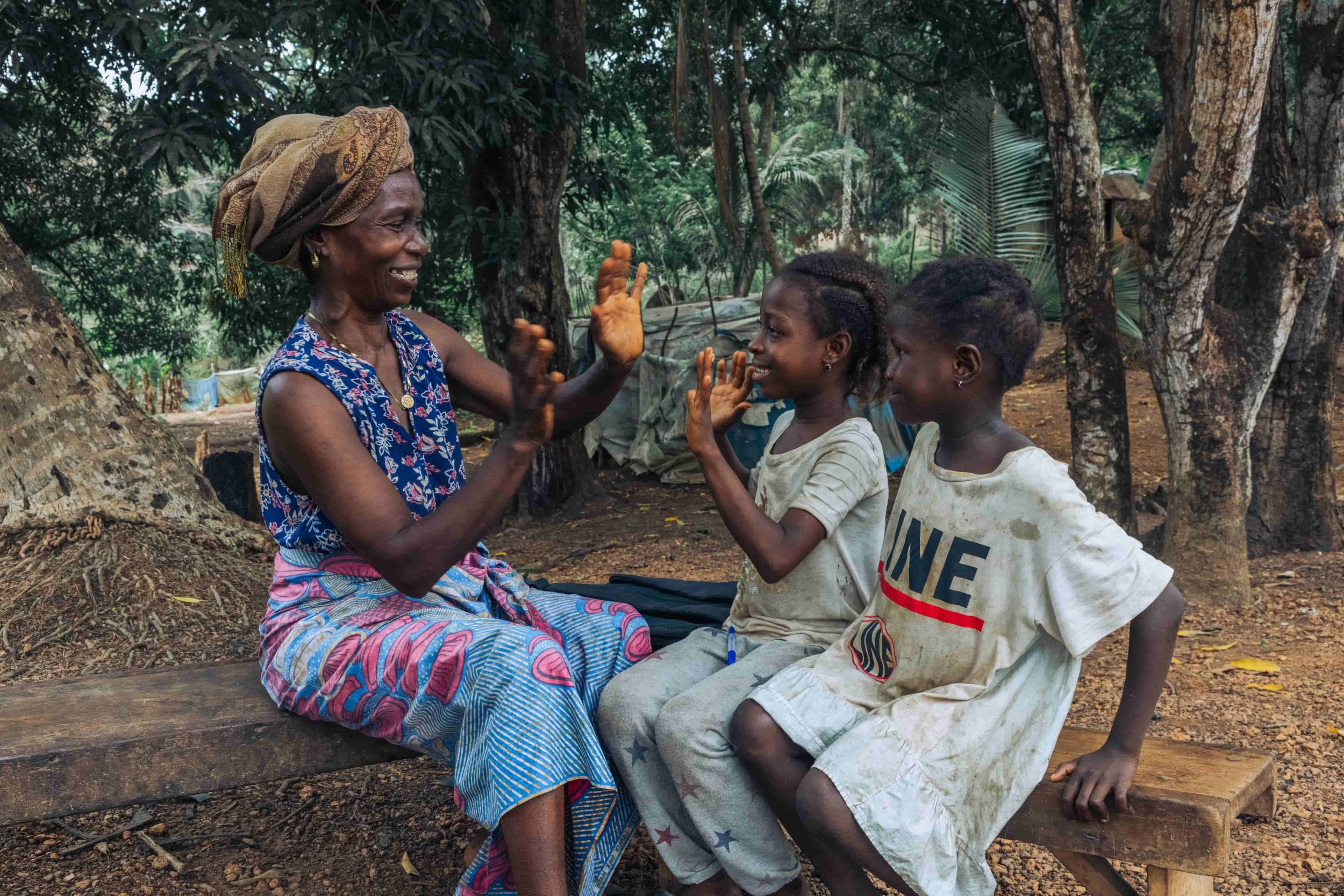 An elderly woman with a headwrap and traditional dress sitting on a wooden bench, smiling and raising her hand, while two young girls with dark hair and casual clothes sit in front of her, in a forest setting with trees and green foliage in the backg