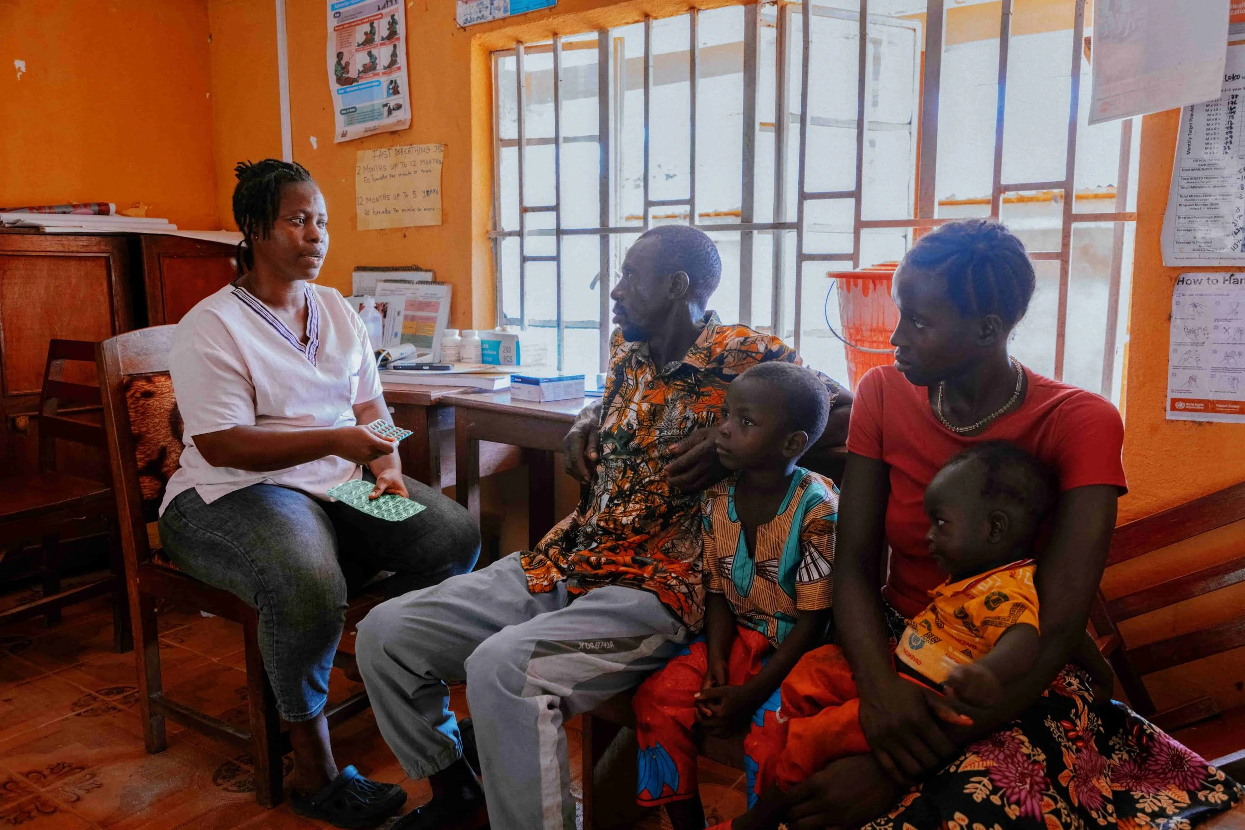A healthcare professional sitting on a wooden chair, talking to a group of four people, including children and adults, inside a room with orange walls and posters on the walls. There is a window with bars behind them.