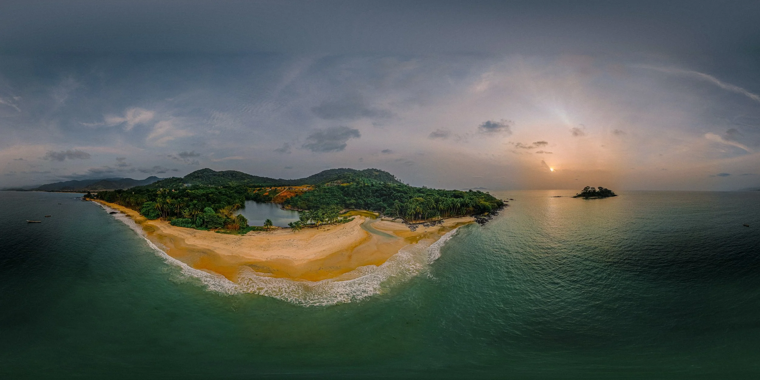 Aerial view of a tropical beach with golden sand, turquoise water, lush green trees, and hills in the background during sunset.