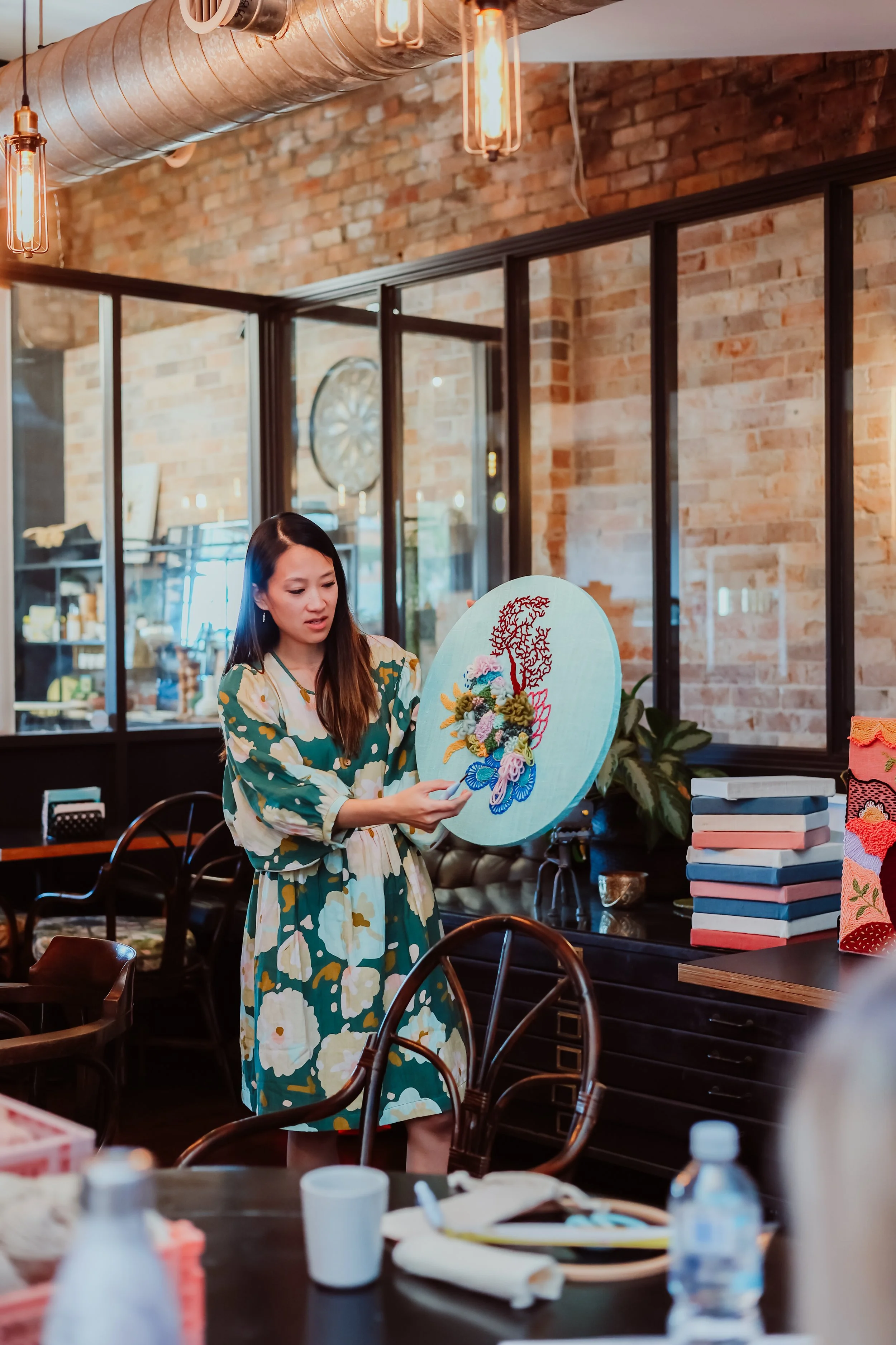 A woman in a floral dress stands in a cafe or restaurant, holding a decorative embroidered piece with colorful floral and butterfly designs. The setting has a rustic brick wall, large window, and warm industrial-style lighting.