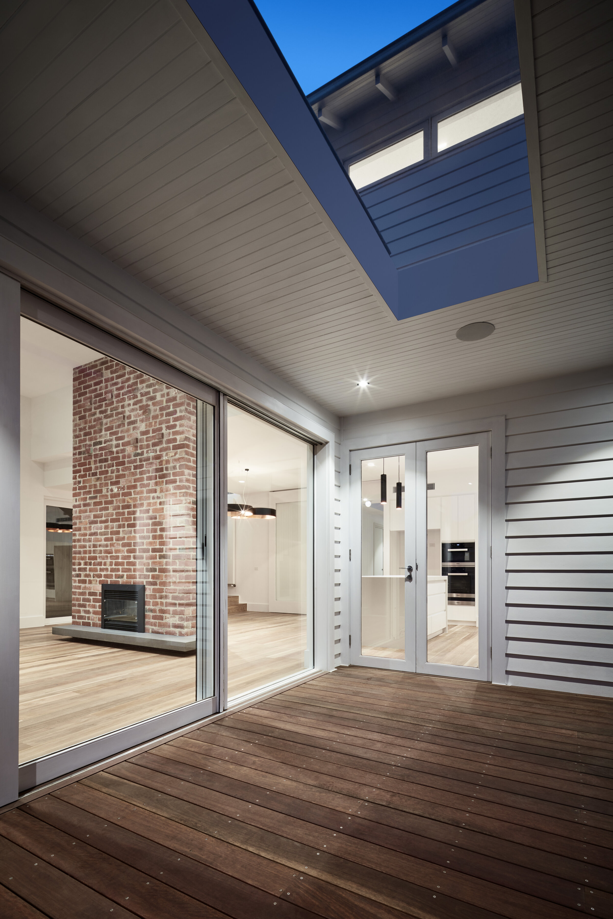 View of a modern covered outdoor patio with wooden decking, sliding glass doors, and an interior living space with a brick fireplace and kitchen.