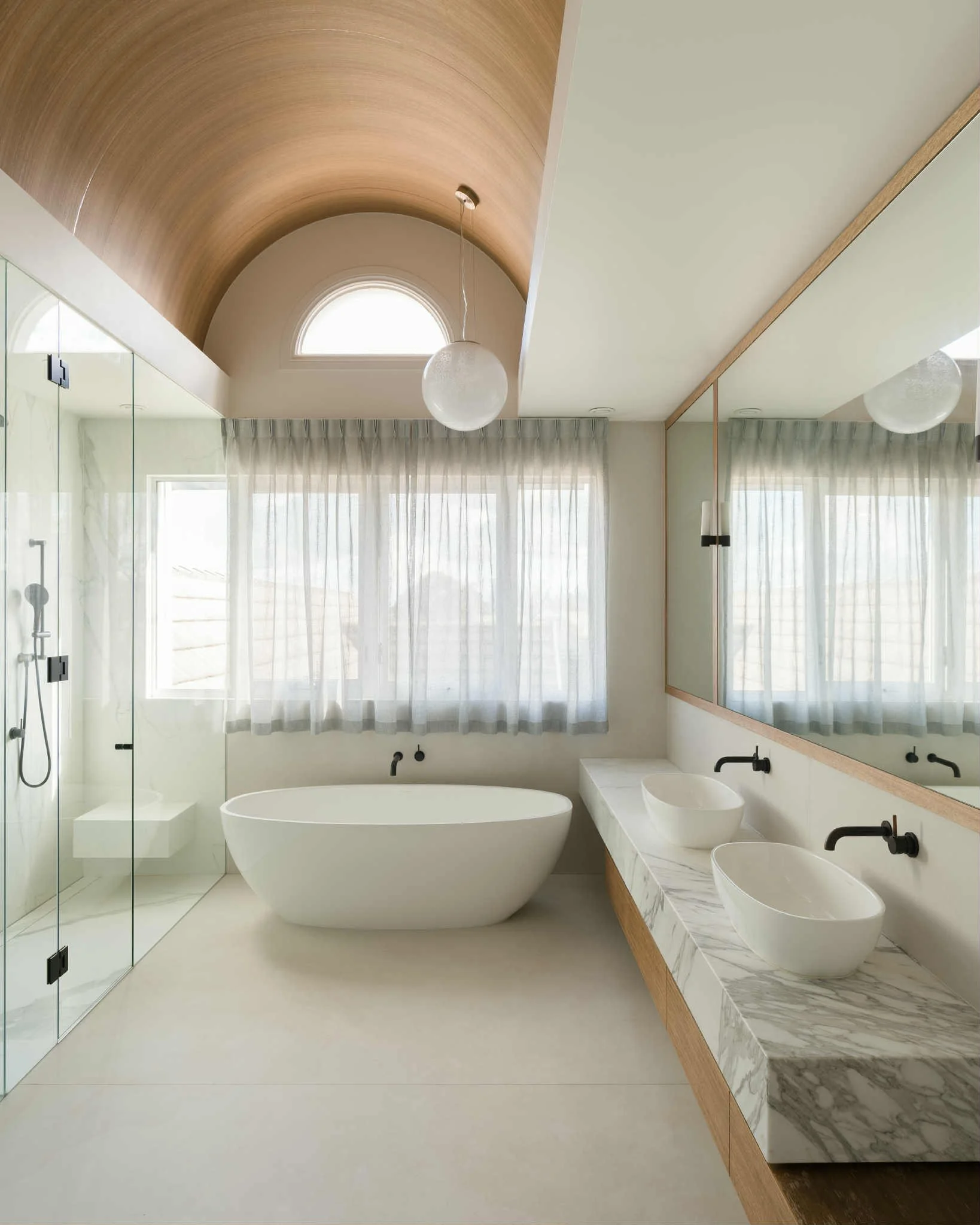 Modern bathroom with a freestanding bathtub, two vessel sinks on a marble countertop, a large mirror, and a walk-in shower with glass door. Natural light through windows with sheer curtains.