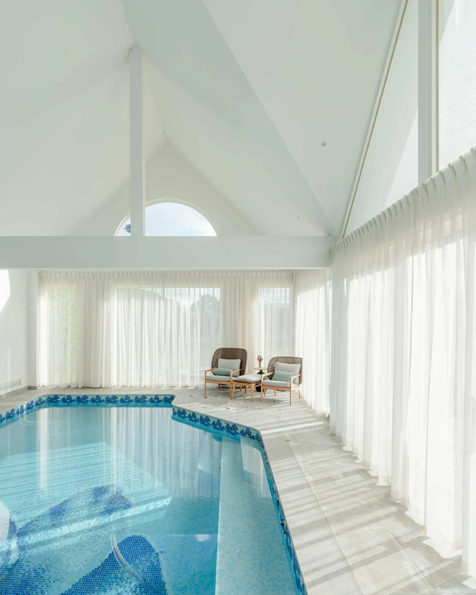 Indoor pool area with large windows and white curtains, two chairs with cushions, and a high ceiling with an arched window.