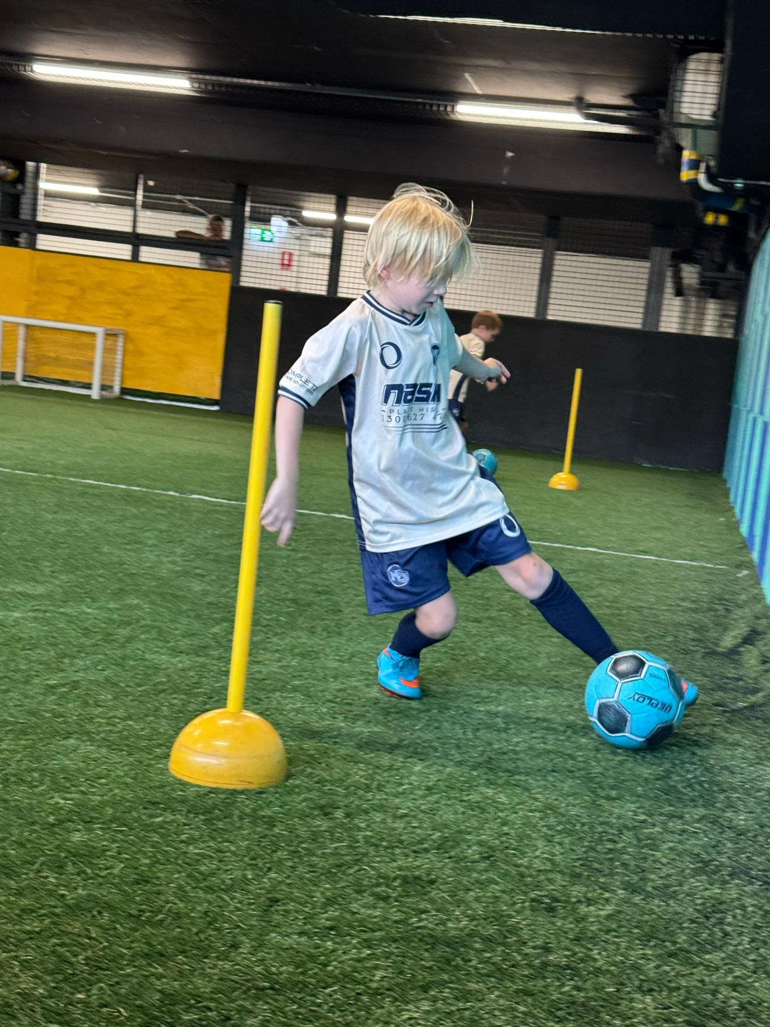 A young boy with blonde hair dribbling a blue and black soccer ball around yellow agility poles at the M5 Centre of Excellence.