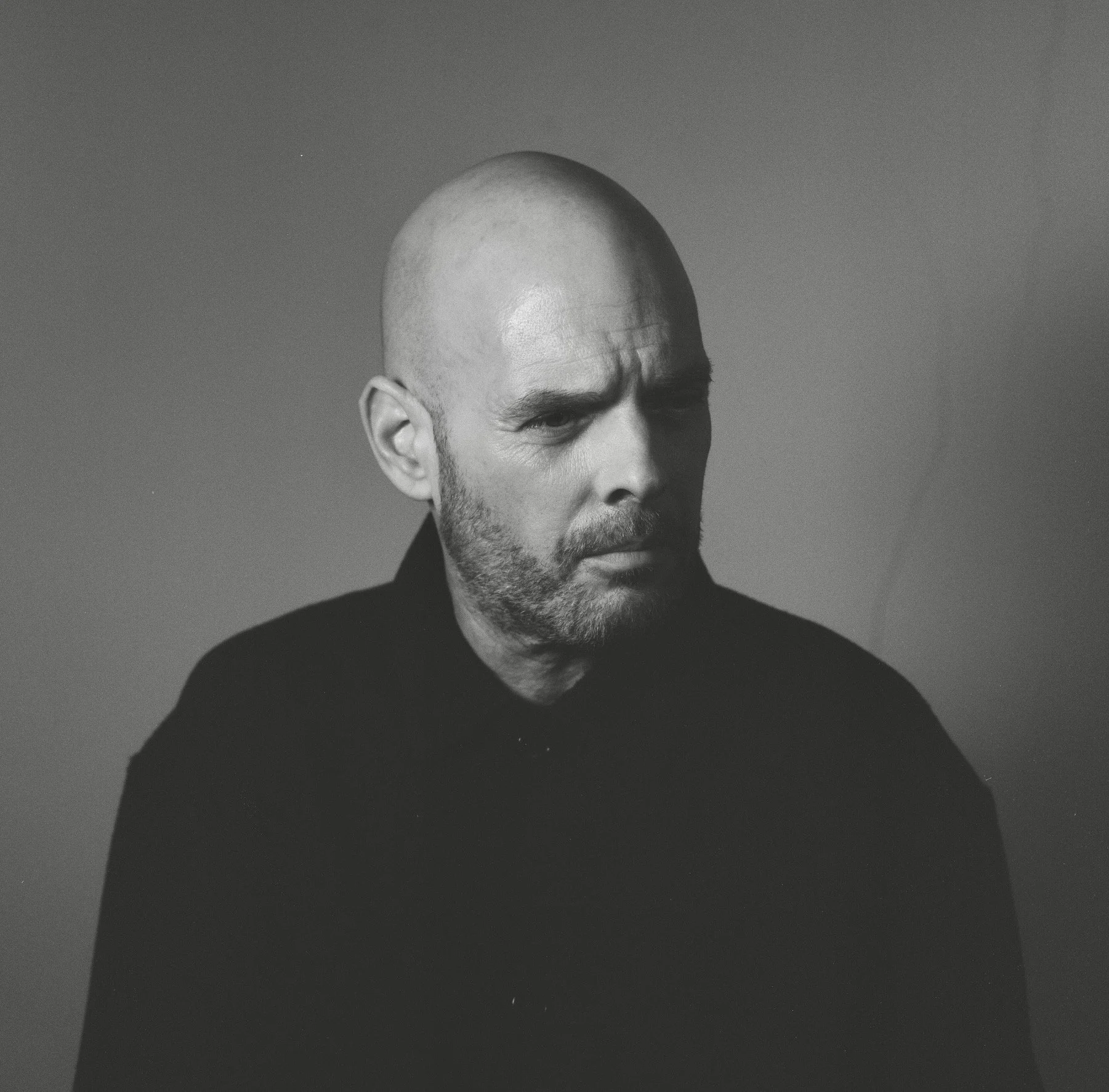 Black and white portrait of a man with a shaved head and beard, wearing a dark shirt, looking downward with a serious expression.