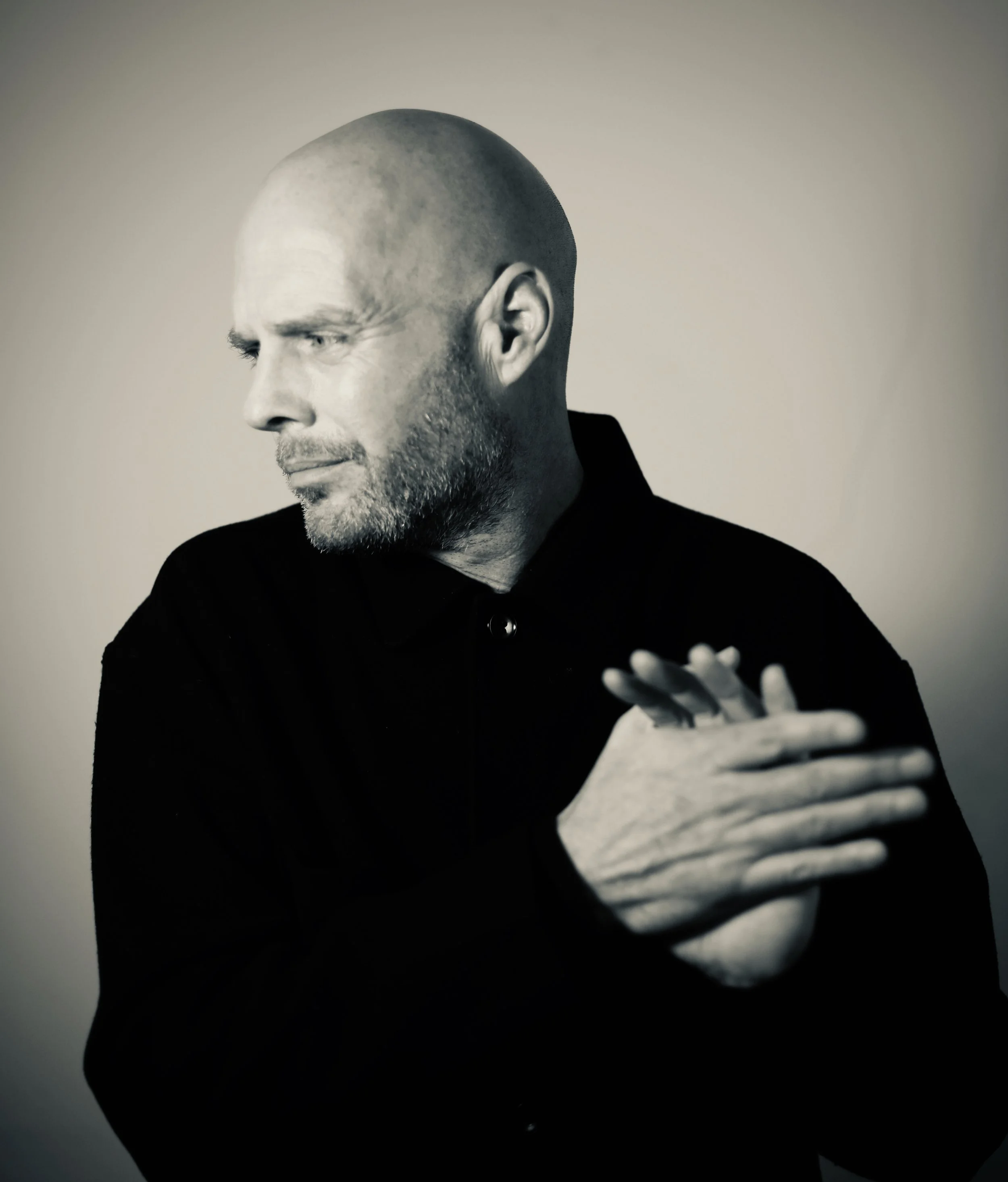 Black and white photo of a bald man with a beard, looking down and to the side, with his arms crossed and wearing a dark jacket.