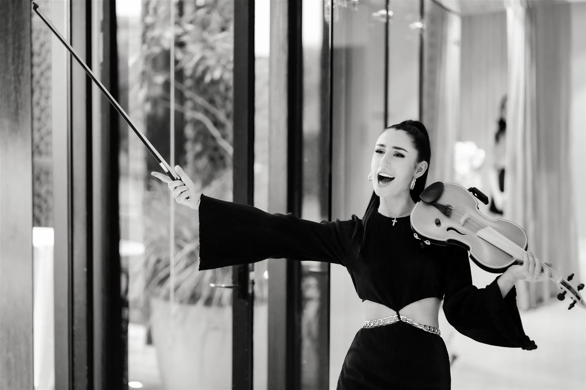 Evangeline Victoria exudes joy and energy, holding her violin and bow triumphantly. Dressed in a stylish black ensemble with gold accents, she shines in this dynamic black-and-white shot, embodying her passion for performance.