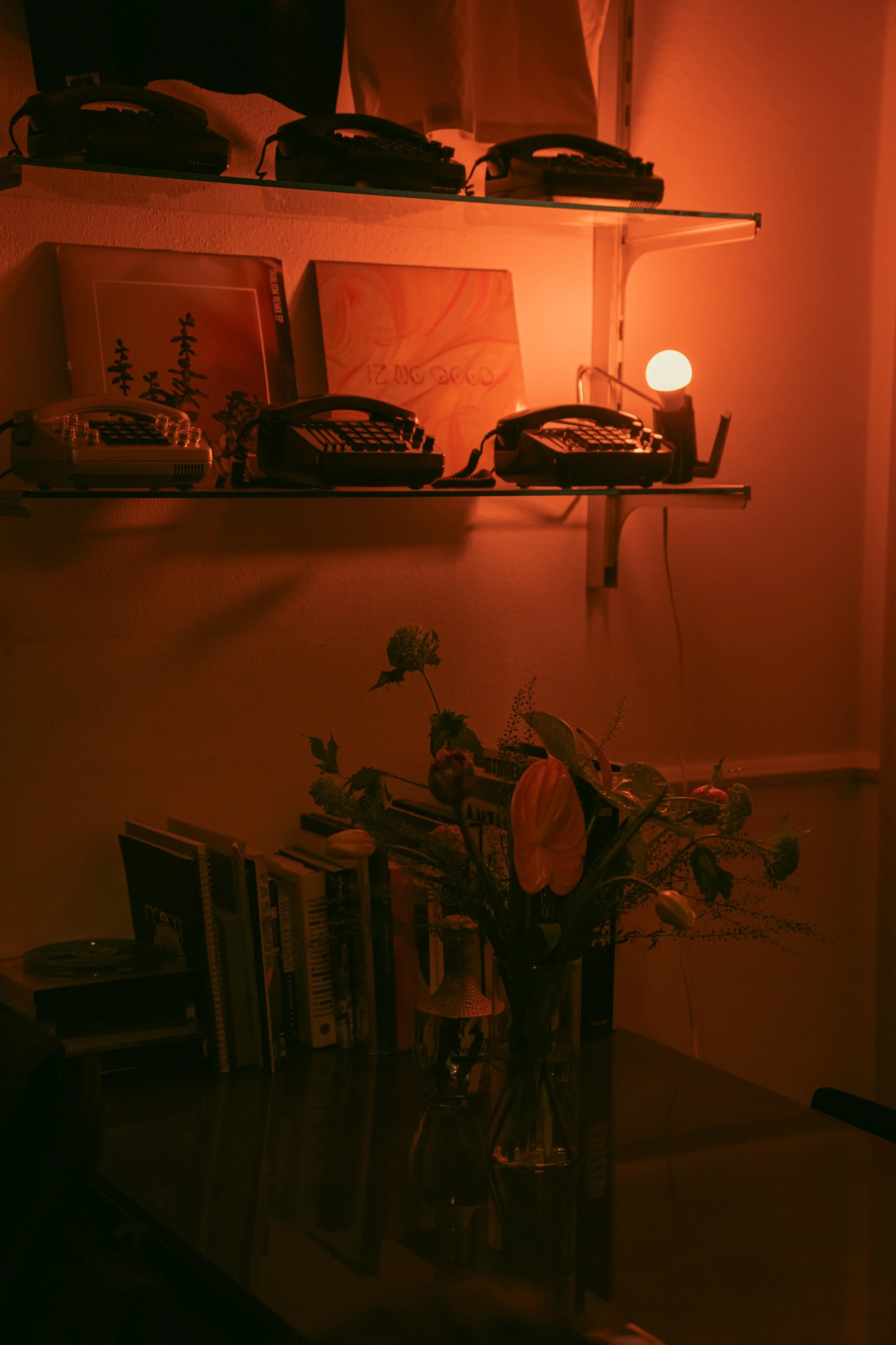 A room with two black shelves on a wall, holding vintage corded phones. There are two picture frames on the lower shelf, one with abstract art and the other with text. Below the shelves, there is a black table with a large flower arrangement in a tall glass vase and a stack of books.
