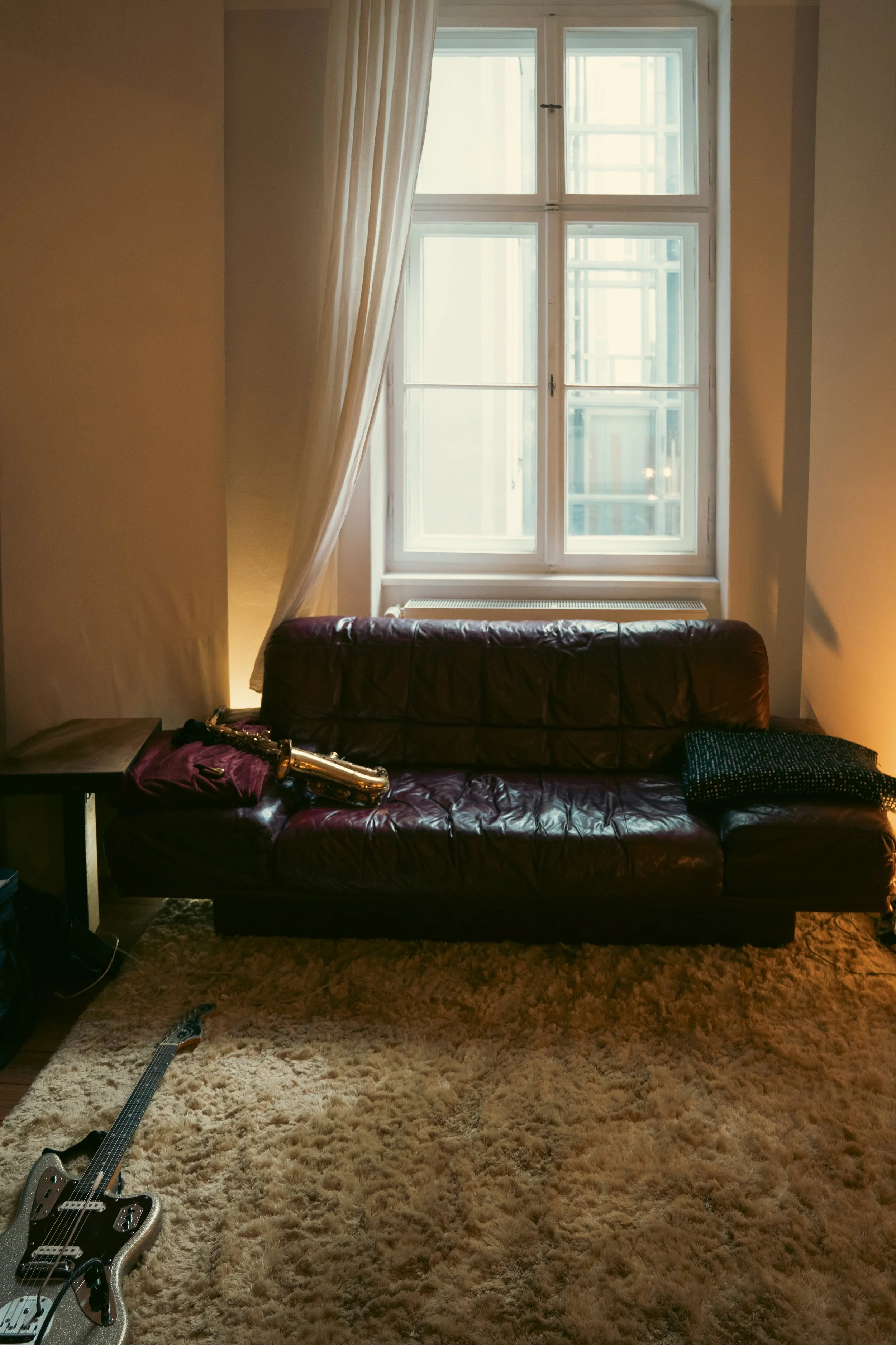 A room with a large window with white framing, a curtain partially drawn, and a dark leather couch in front. On the couch, there is a purple cloth with a shiny gold saxophone and some other small items. To the left, part of a side table and a guitar lying on a thick beige carpet. Sunlight filters through the window.