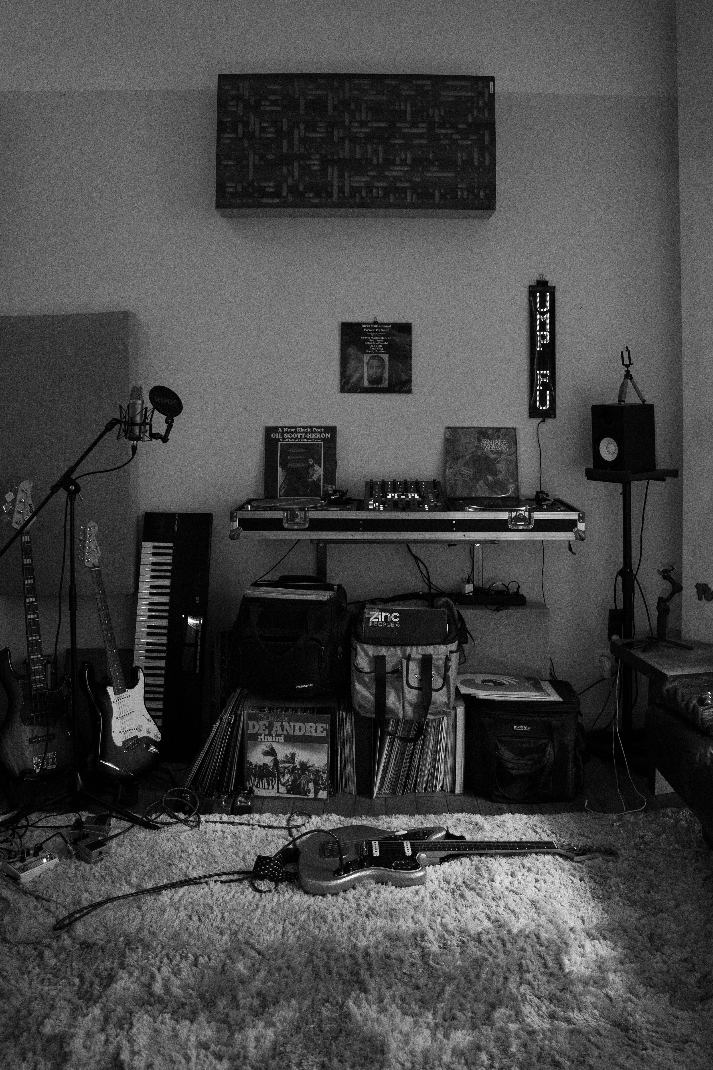 A music studio setup with guitars on the left, a keyboard, vinyl records, and music equipment on shelves. There are posters and a neon sign on the wall that says 'UP'.