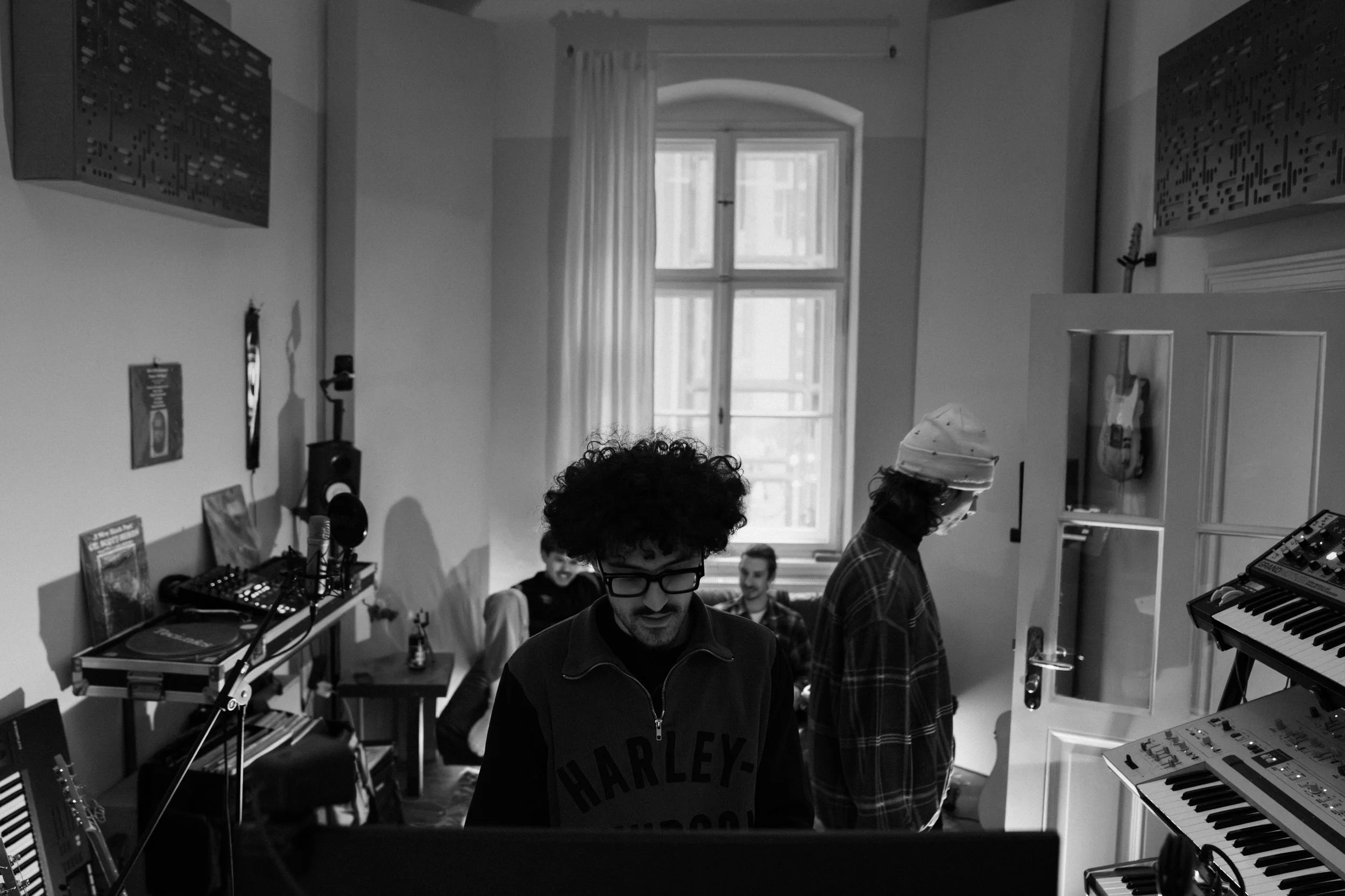 A group of young people in a music studio. Some are standing and some are sitting, surrounded by musical equipment such as keyboards, synthesizers, and audio gear. Large windows with curtains are in the background, with daylight shining through.