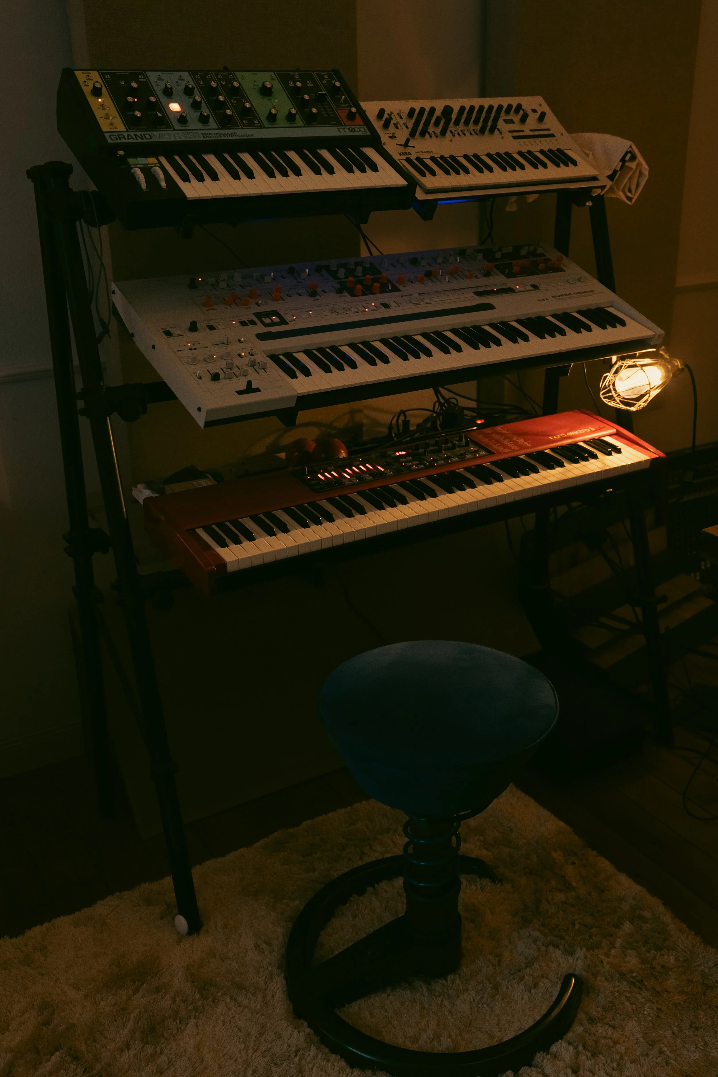 A music studio setup with multiple electronic keyboards and synthesizers on a black multi-tiered stand, illuminated by warm lighting, with a stool in the foreground, a shaggy rug on the floor, and a wall in the background.