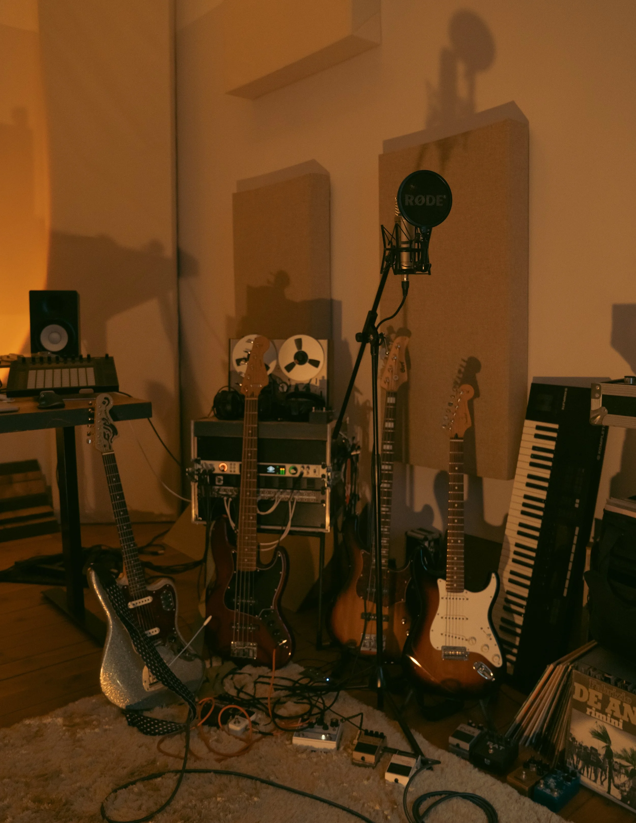 A music studio setup with multiple electric guitars on stands, a keyboard, various pedals on the floor, a microphone on a stand, a small amplifier, and vinyl records on the floor. The room has acoustic panels on the walls.