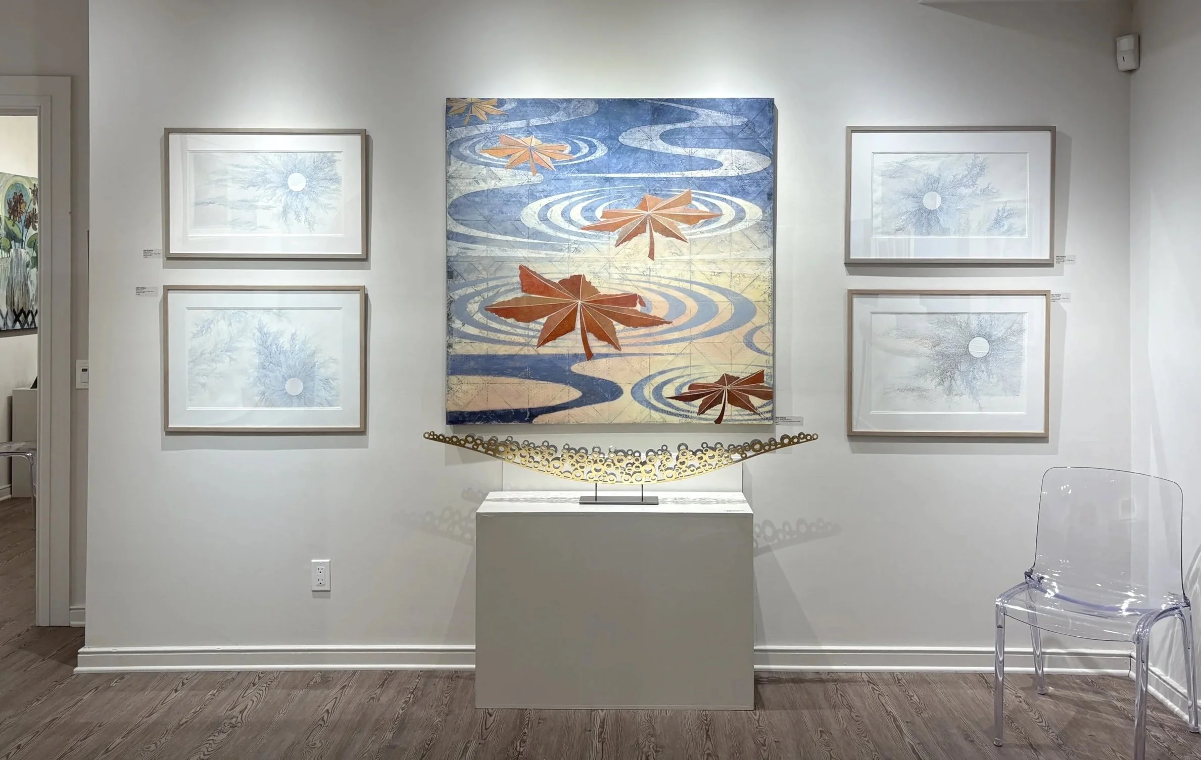 Art gallery wall displaying five pieces of art: a large painting of autumn leaves floating on water with ripples, four smaller framed prints of trees with circular reflections, and a modern circular sculpture on a pedestal at the center, with a clear acrylic chair to the right.