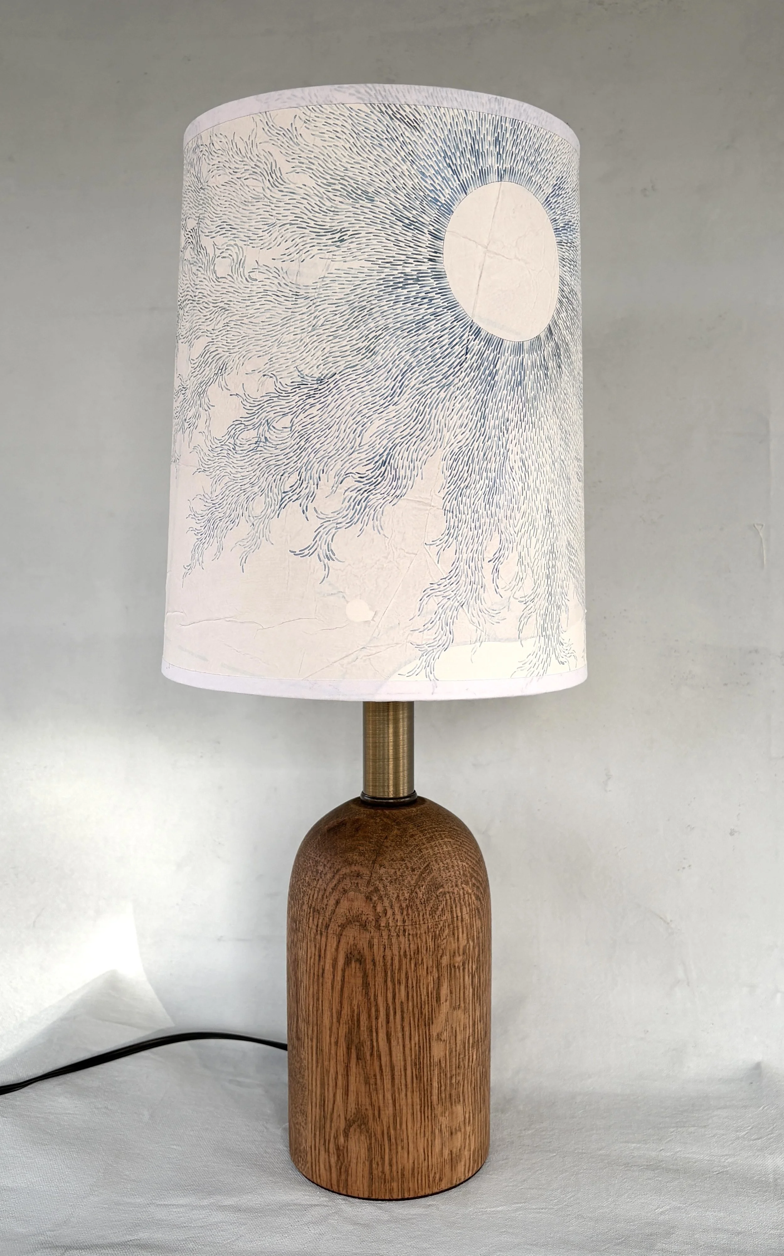 Wooden lamp with cylindrical base and a white shade featuring a simple line drawing of a circular shape with radiating lines, resembling a sun or moon.