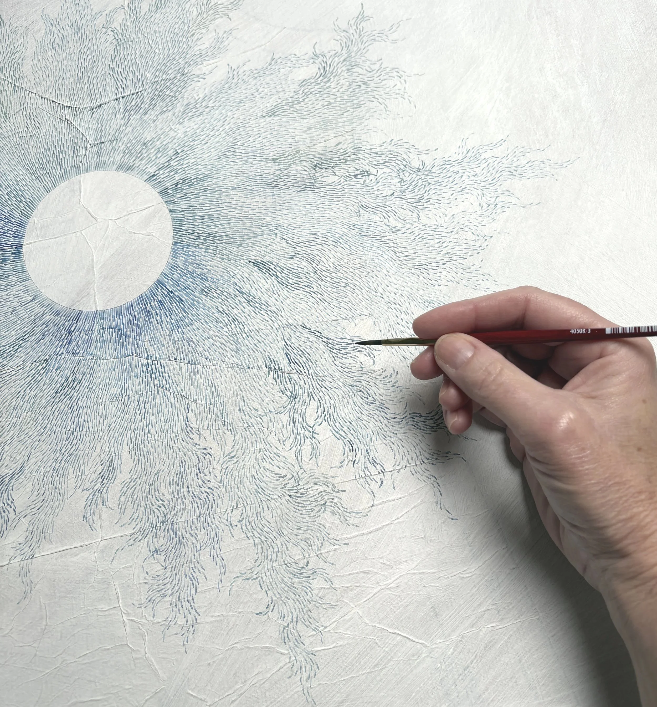 An artist's hand is creating a detailed, abstract illustration on textured white paper with a fine-tipped brush. The artwork features numerous fine lines forming a circular pattern with radiating wisps and a central circle.