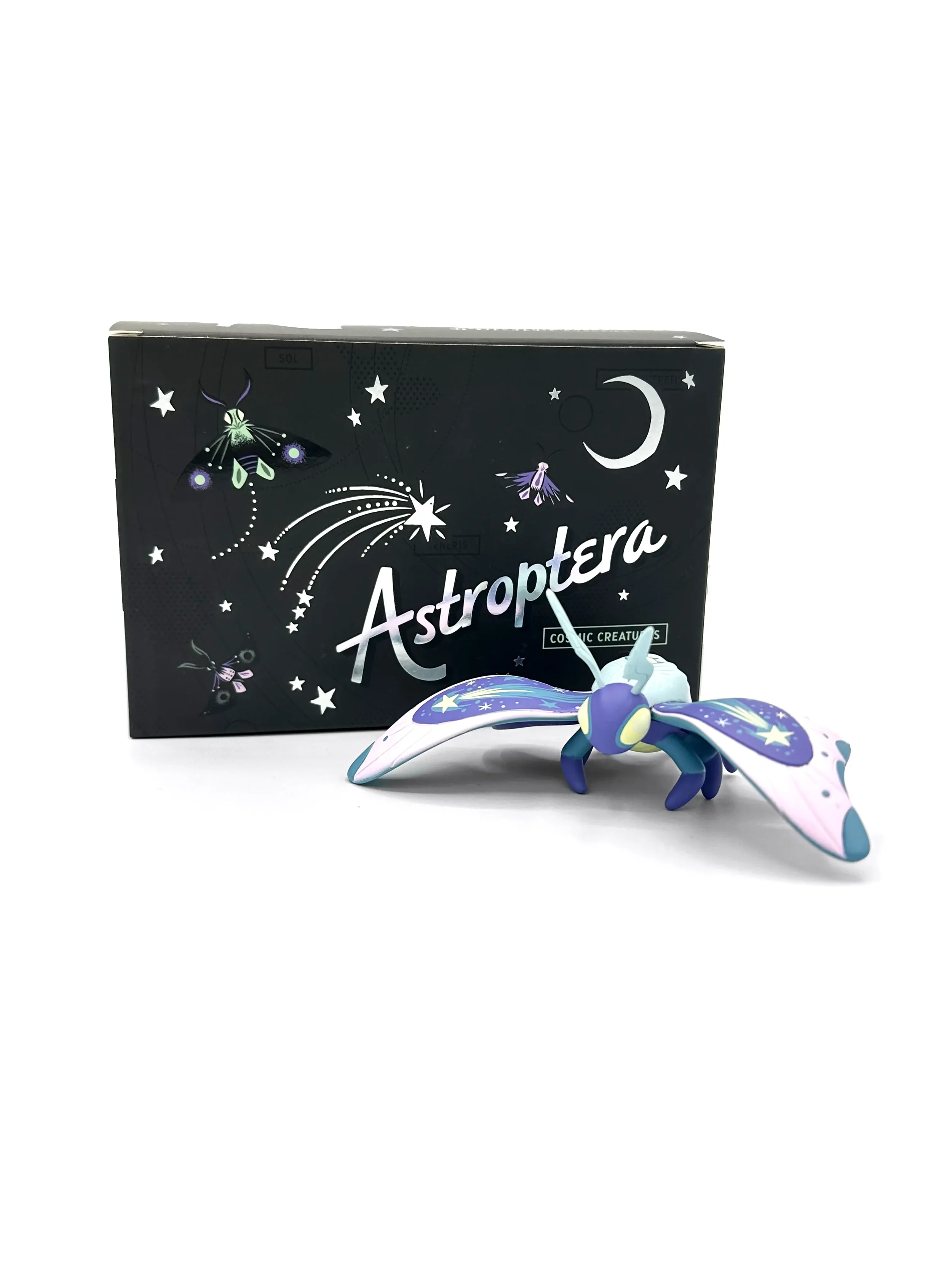 Stephanie Buscema Astroptera Stelle vinyl moth art toy with wings spread beside Cosmic Creatures collector box