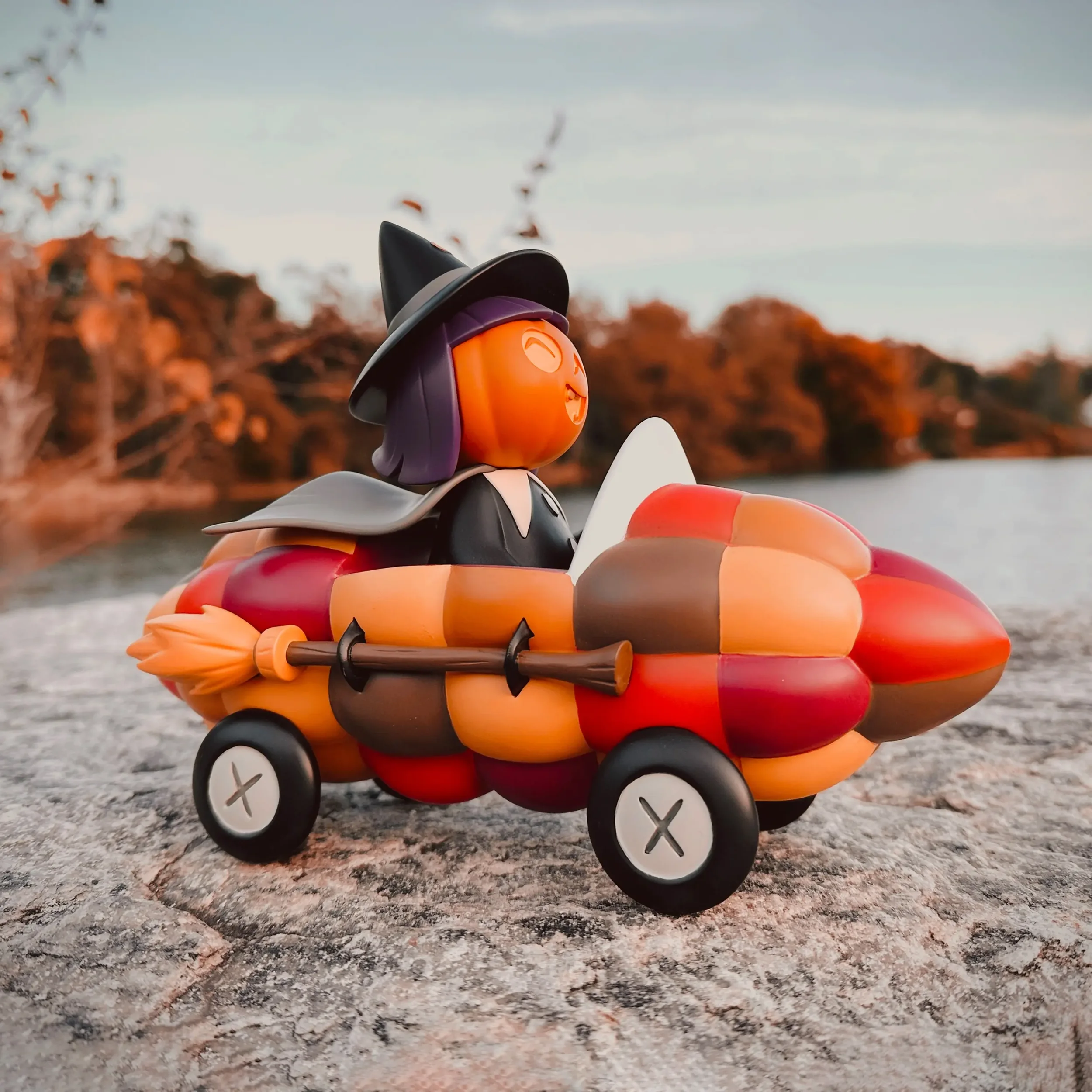 Stephanie Buscema Willa the Way-Out Witch resin art toy on stone ledge at sunset with autumn lake and foliage background