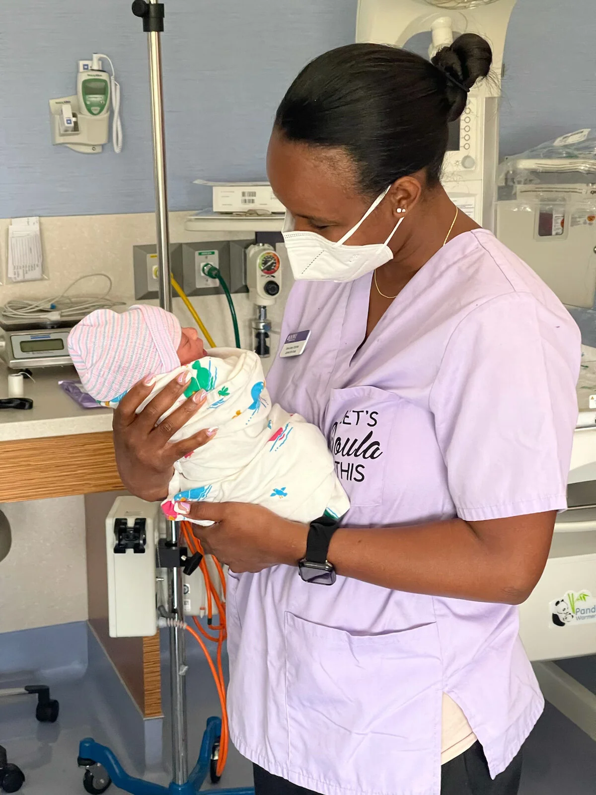 A nurse in a pink uniform and face mask holding a newborn baby wrapped in a blanket, in a hospital setting.