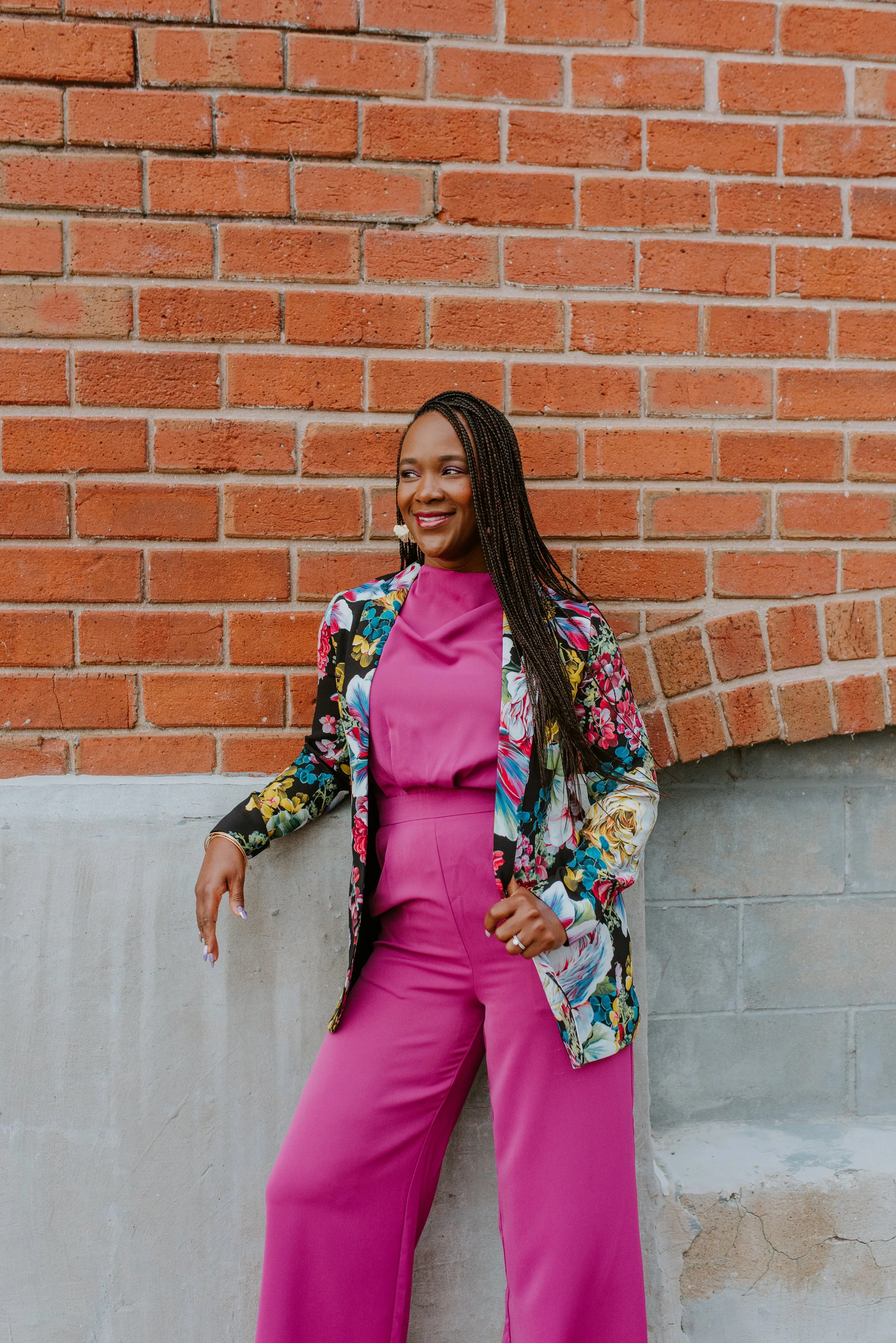 A woman with long braided hair wearing a bright pink jumpsuit and a multicolored floral blazer standing outdoors against a brick and concrete wall, smiling and looking to her right.